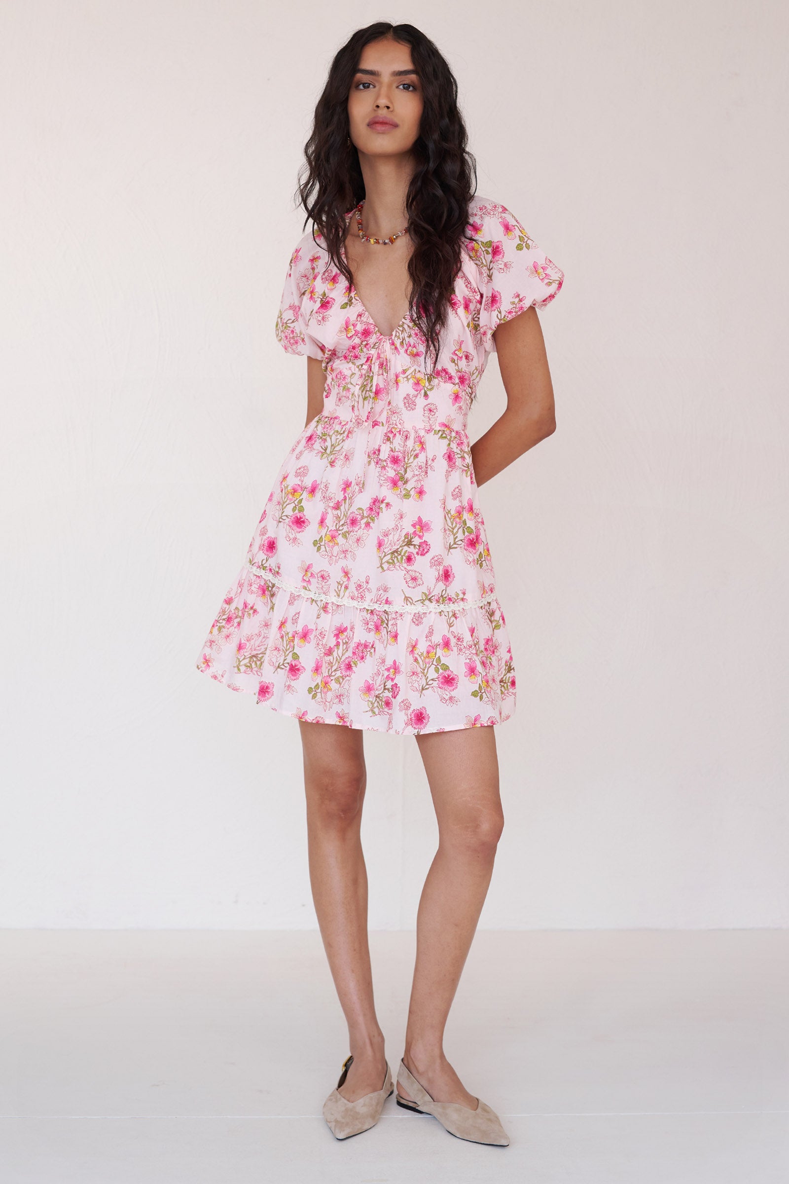 Front view of a woman standing wearing the Artemis dress in Burdock floral print in Peony color. It features a drawstring neckline, ladder lace detailing, elasticated sleeves, princess-seamed waist & side-seam pockets.