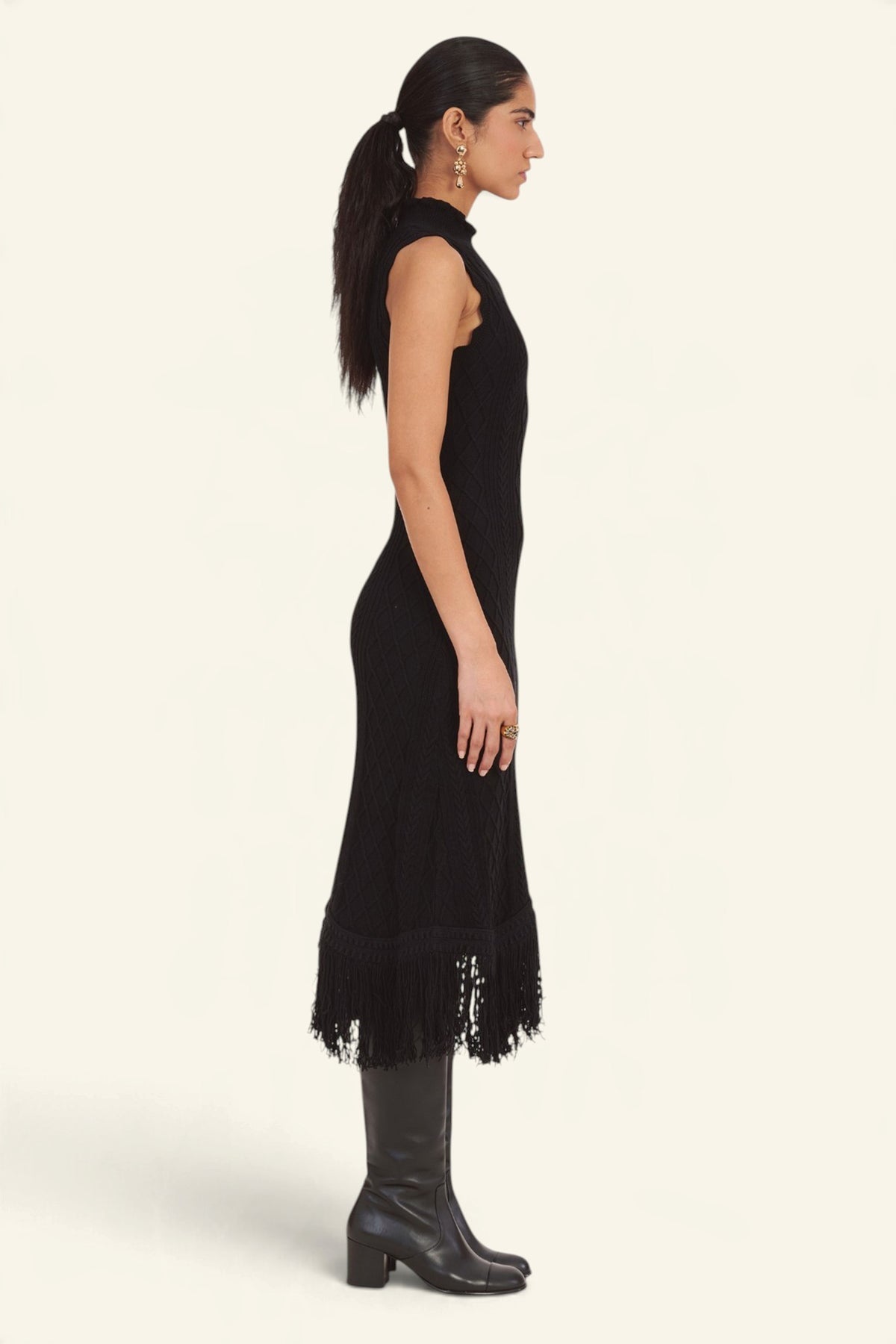 Side view of model standing wearing the Amelia Fringe Dress, featuring a body-skimming cable-knit silhouette, high neckline with hand-crocheted trim, and an uneven hem finished with fringe.