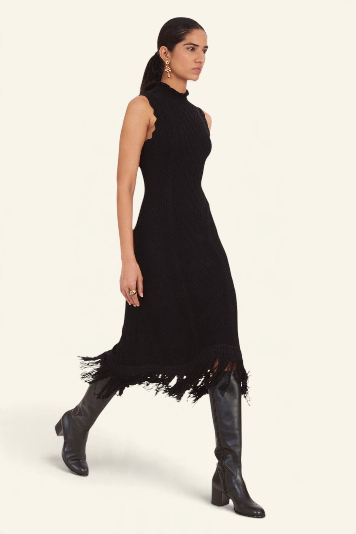 Side view of model walking wearing the Amelia Fringe Dress, featuring a body-skimming cable-knit silhouette, high neckline with hand-crocheted trim, and an uneven hem finished with fringe.