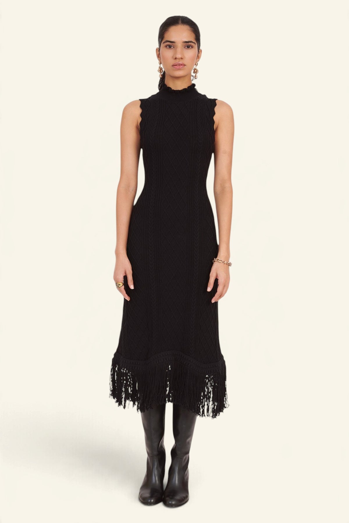 Front view of model standing wearing the Amelia Fringe Dress, featuring a body-skimming cable-knit silhouette, high neckline with hand-crocheted trim, and an uneven hem finished with fringe.