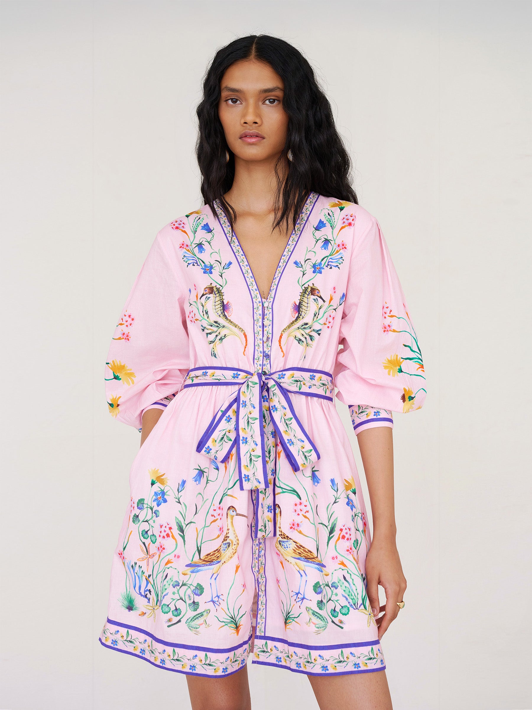 Front view of a woman standing. She is wearing the Anne mini dress in Tidepool print conch color with a hand in pocket. It features long sleeves, a short hemline, an elasticated waist, fabric-covered buttons, and a fabric belt.