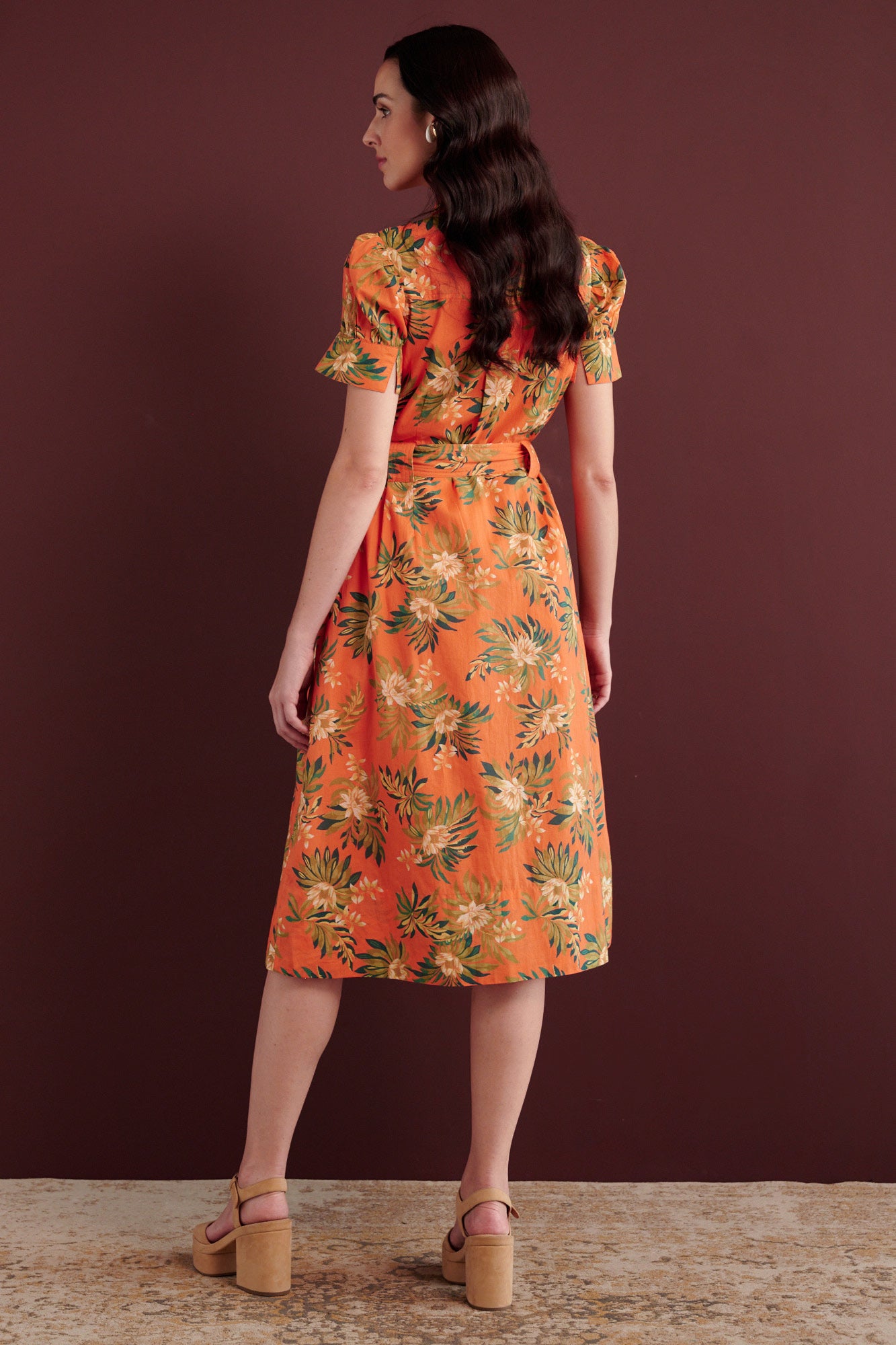 Back view of woman standing. She is wearing the Barbara day dress in Artichoke Flower print. It has gathered sleeves, chest pockets, fabric belt, button details, and a subtly structured yet romantic silhouette.