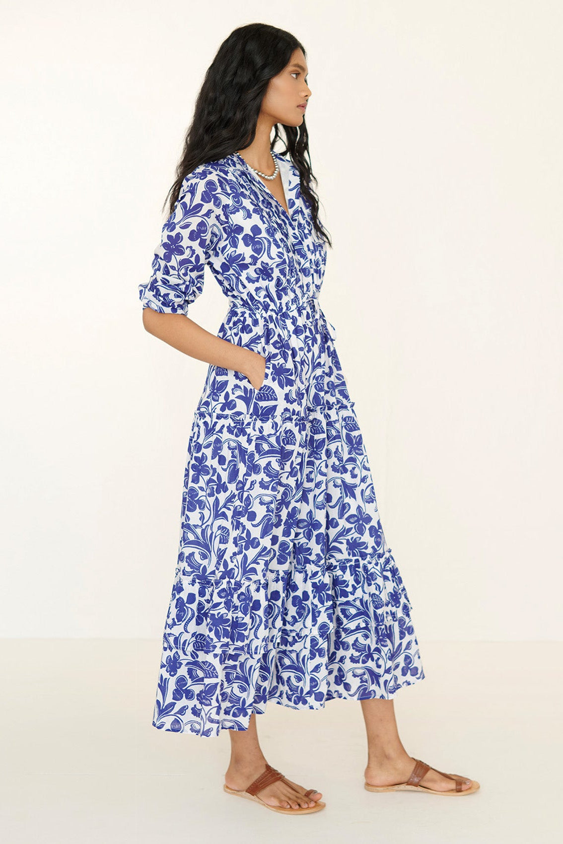 Side view of woman wearing Bazaar Dress in blue and white Gaia floral print cotton voile with long sleeves, tiered ankle-length skirt, drawstring belt, and side pockets.