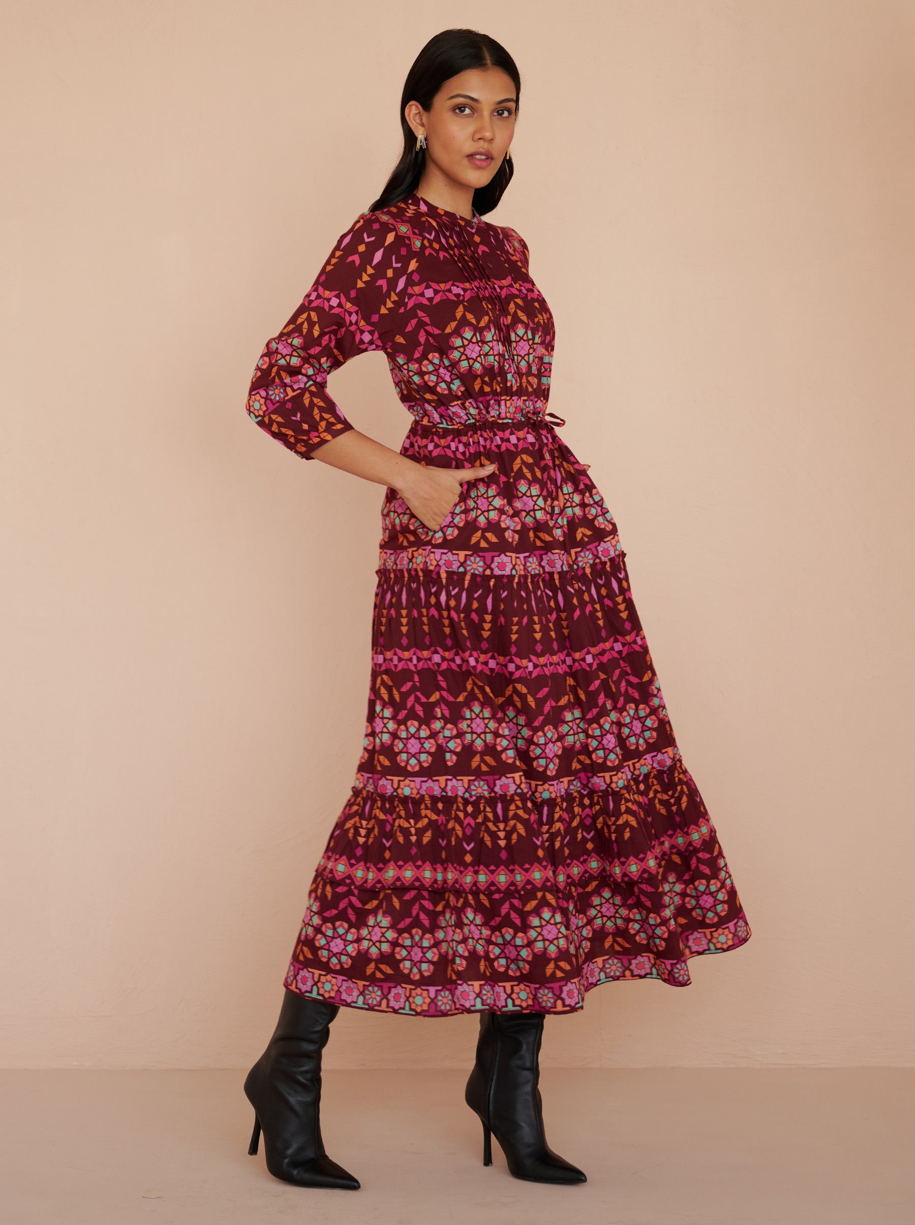 Side view of a model standing with hands in pocket. She is wearing the Bazaar ankle-length dress in cotton voile with the Tapestry print in Rhubarb color. It is featuring long sleeves, a tiered A-line skirt, a drawstring self-belt, full lining, and side-seam pockets.