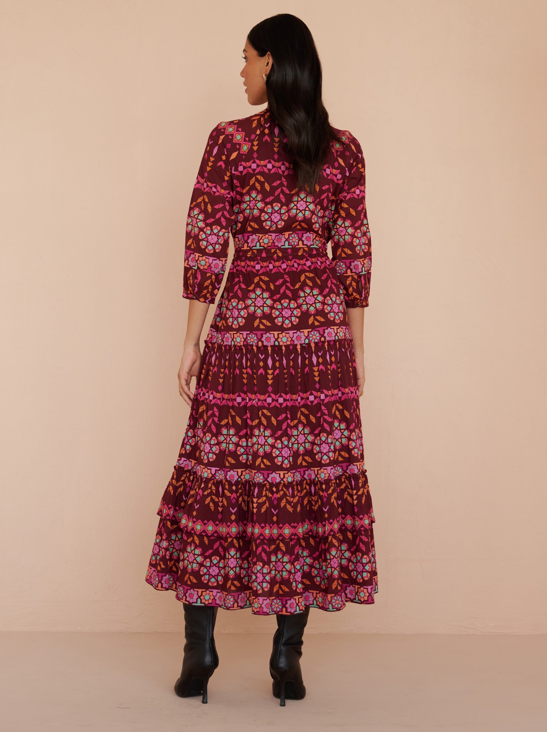 Back view of a model standing. She is wearing the Bazaar ankle-length dress in cotton voile with the Tapestry print in Rhubarb color. It is featuring long sleeves, a tiered A-line skirt, a drawstring self-belt, full lining, and side-seam pockets.