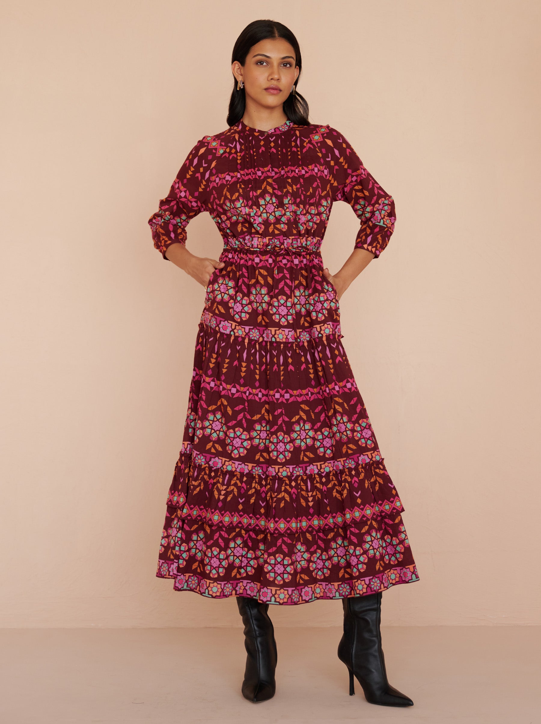 Front view of a model standing. She is wearing the Bazaar ankle-length dress in cotton voile with the Tapestry print in Rhubarb color. It is featuring long sleeves, a tiered A-line skirt, a drawstring self-belt, full lining, and side-seam pockets.