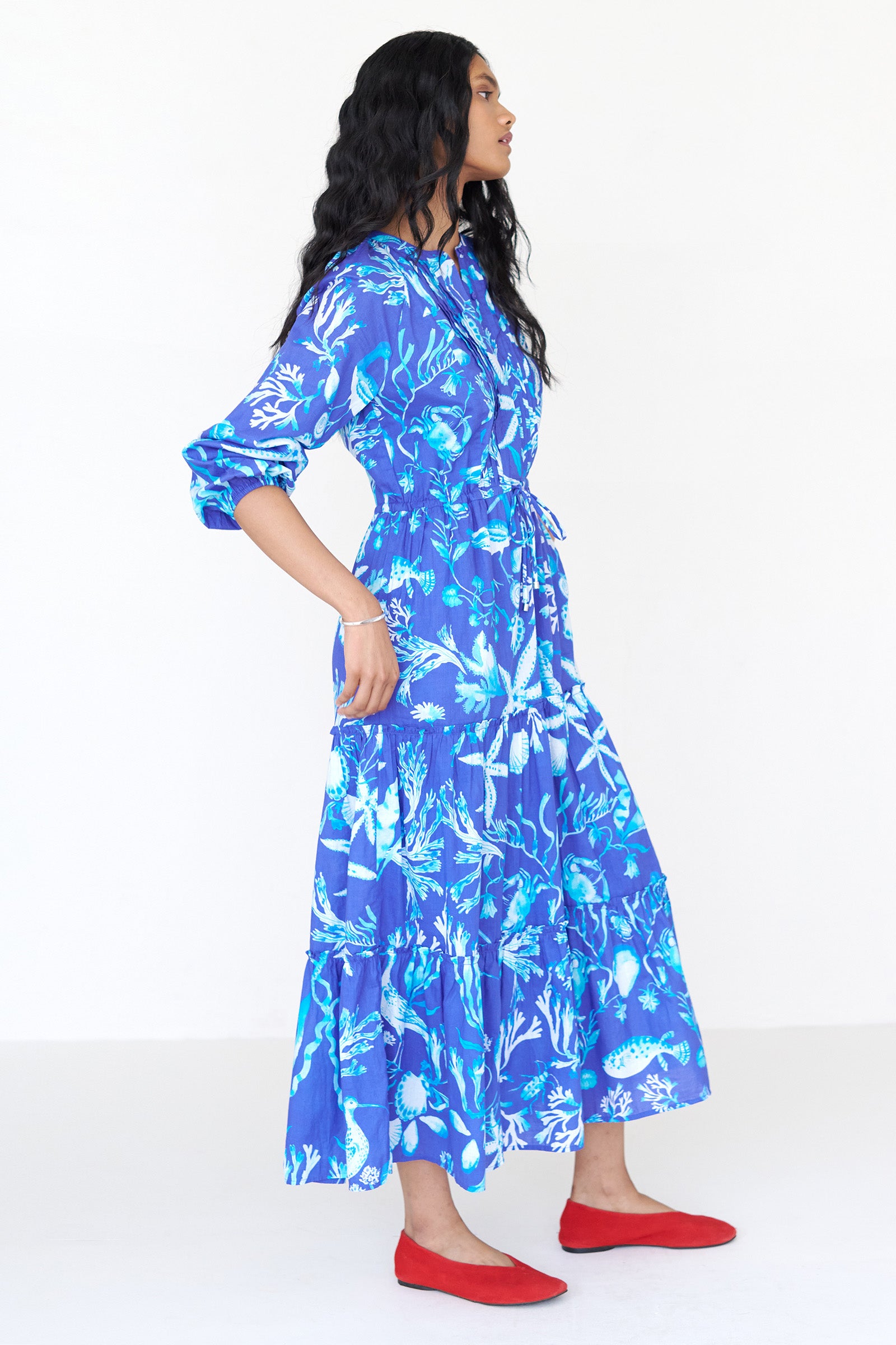 Side view of a woman standing wearing the Bazaar dress in Tidepool print. It features long sleeves, tiered ankle-length skirt, drawstring belt and side-seam pockets.