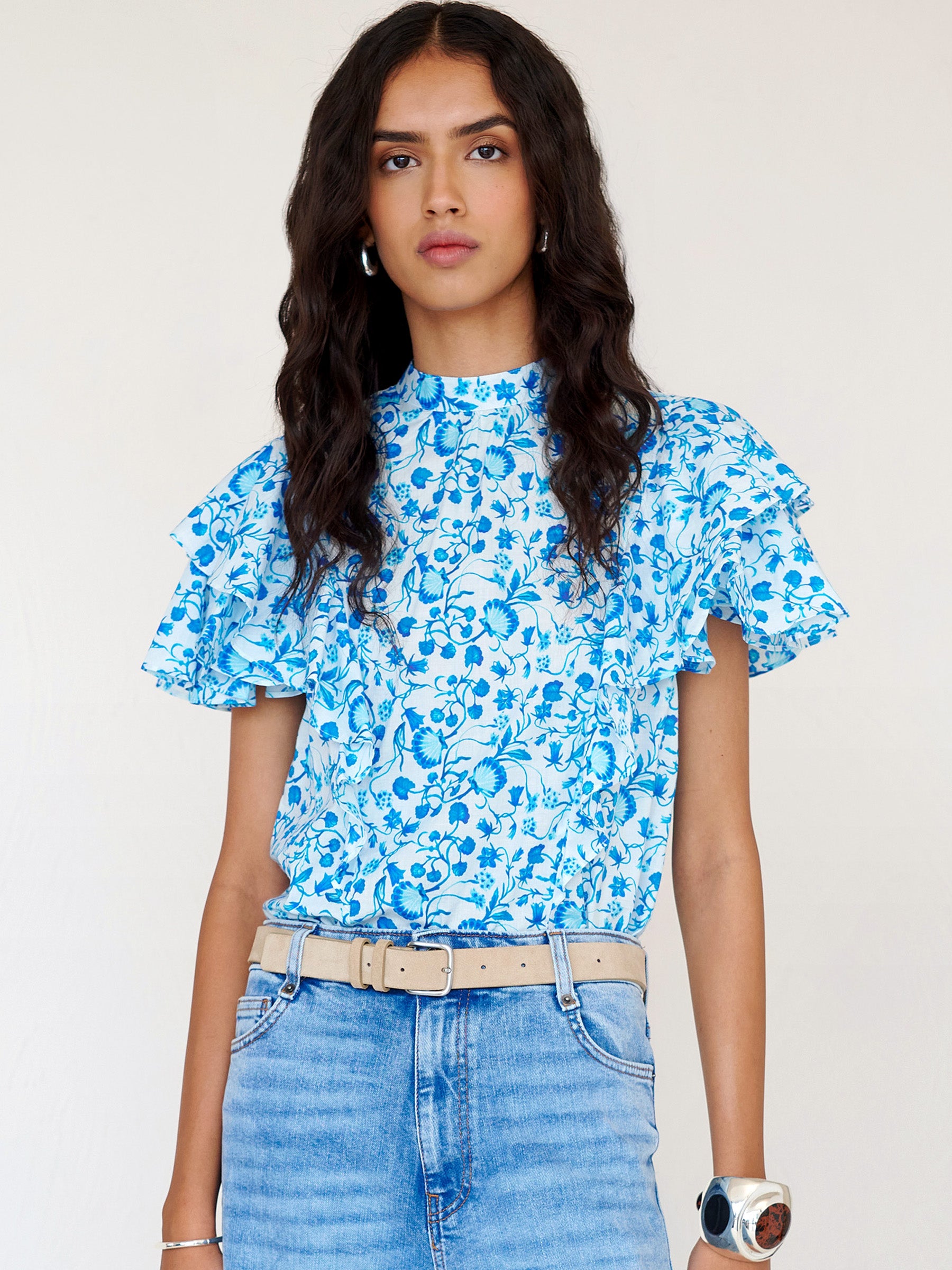 Front view of a woman standing. She is wearing the Bebe top in Clam Garland print paired with denim. It features a soft drape over the shoulder, tie at the neck, and full layered sleeves.