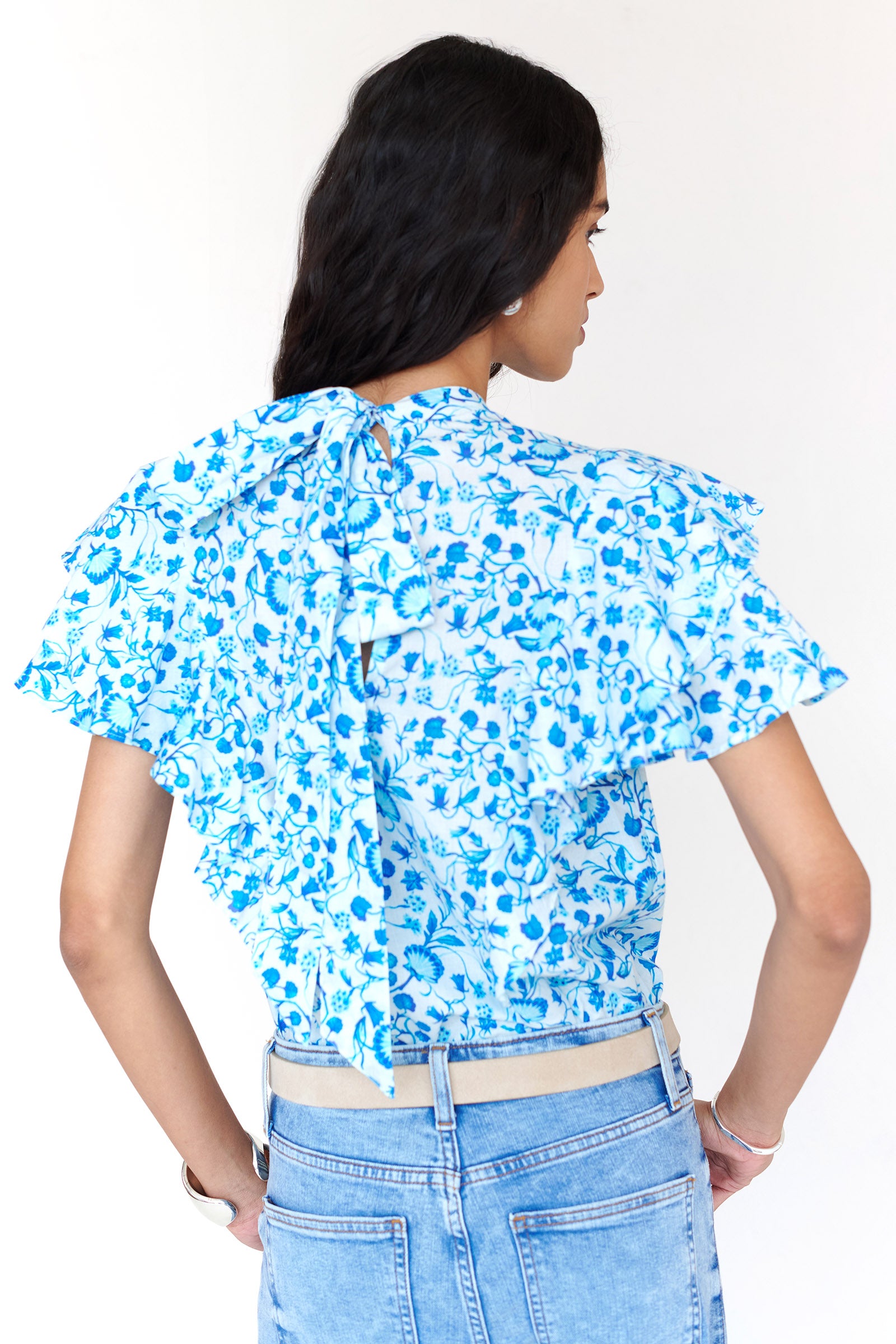 Back view of a woman standing. She is wearing the Bebe top in Clam Garland print. It features a soft shoulder drape, tie neckline, and layered sleeves. Crafted in a timeless floral inspired by the Scottish Isles, styled with denim.