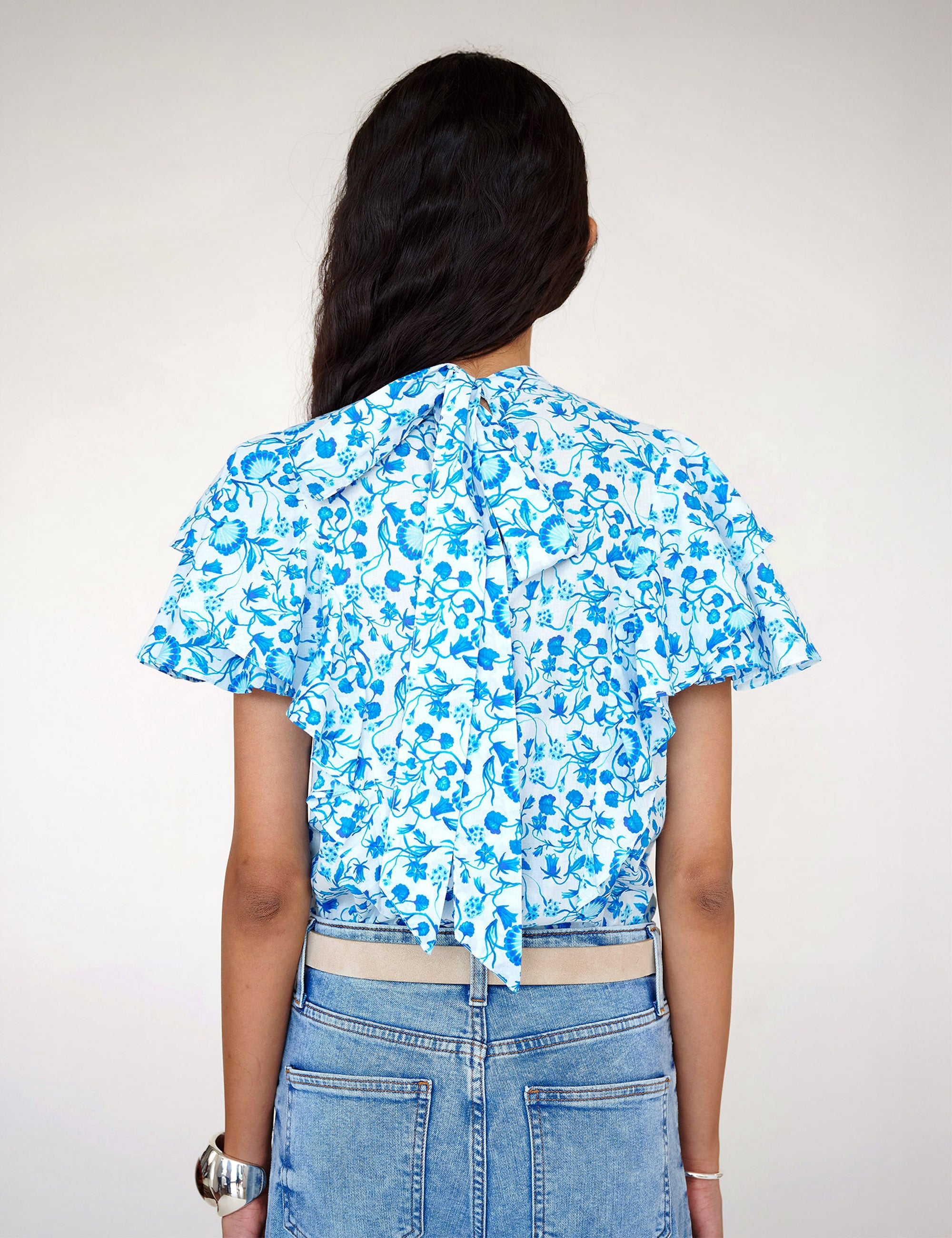Back view of a woman standing. She is wearing the Bebe top in Clam Garland print paired with denim. It features a soft drape over the shoulder, tie at the neck, and full layered sleeves.