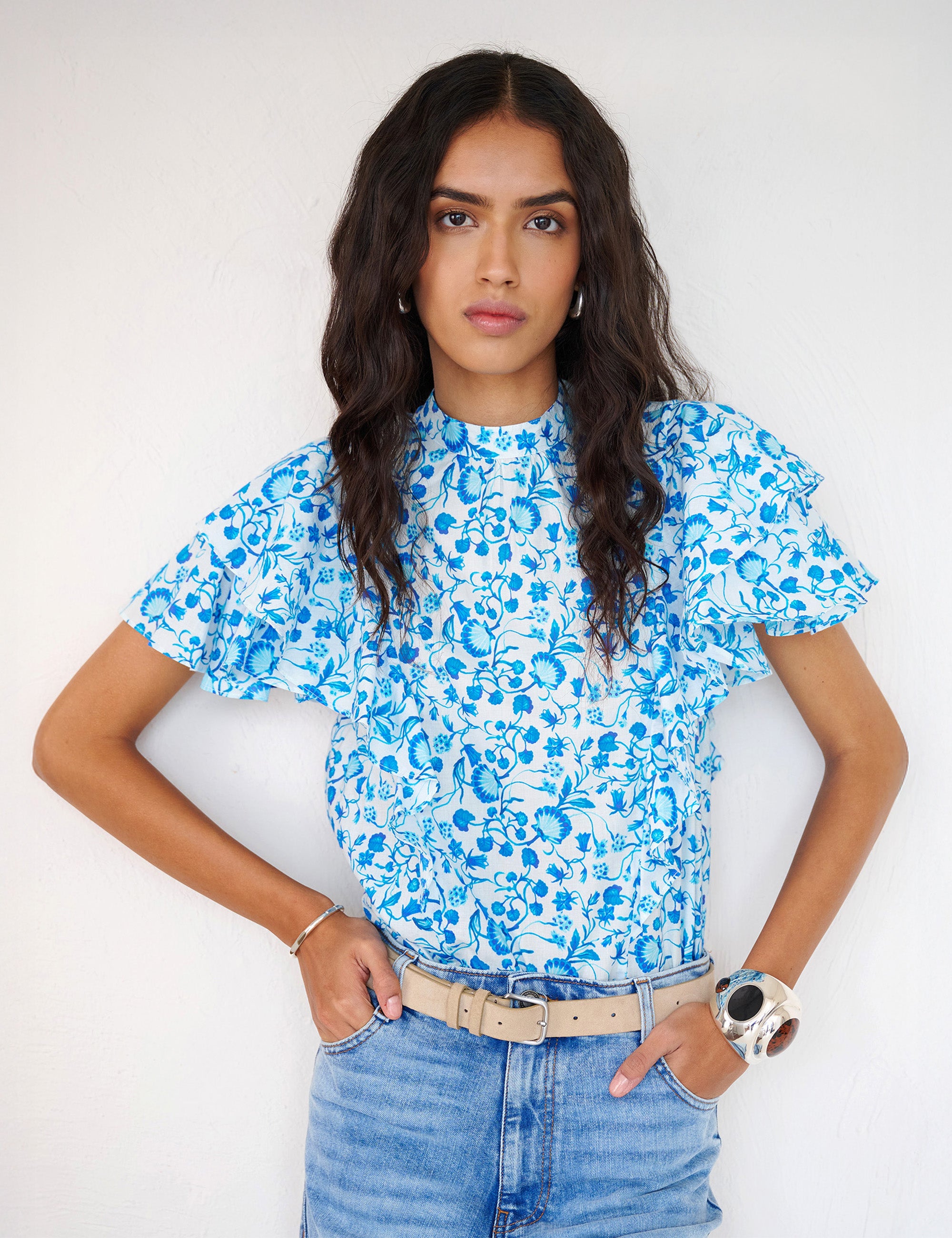 Front view of a woman standing. She is wearing the Bebe top in Clam Garland print paired with denim. It features a soft drape over the shoulder, tie at the neck, and full layered sleeves.