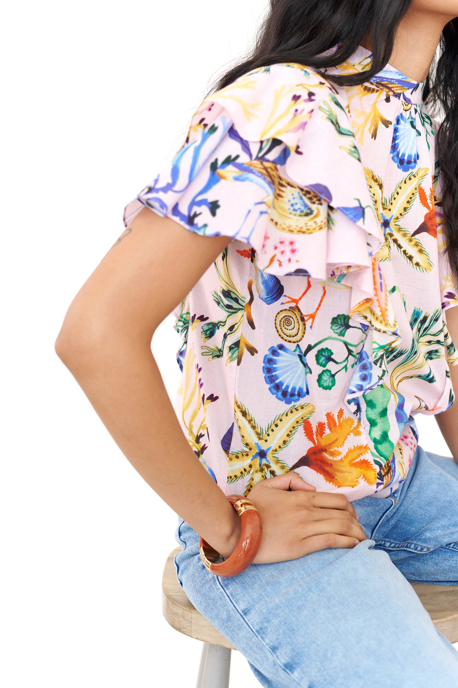 Side view close up of a woman sitting. She is wearing the Bebe top in Tidepool print, featuring a soft shoulder drape, tie neckline, and full layered sleeves. Ocean-inspired print, styled with denim.