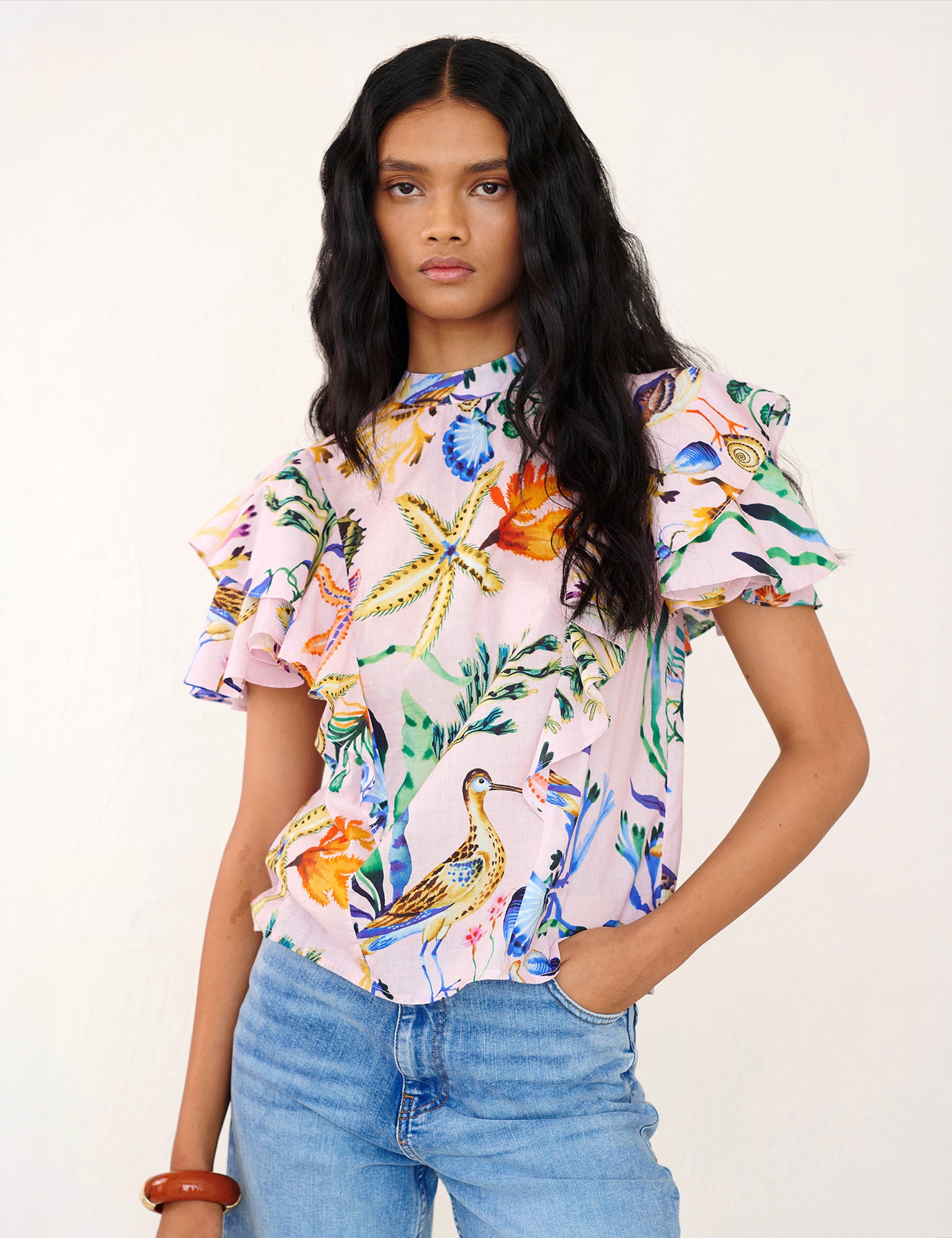 Front view of a woman standing. She is wearing the Bebe top in Tidepool print. It features a soft drape over the shoulder, tie at the neck, and full layered sleeves.