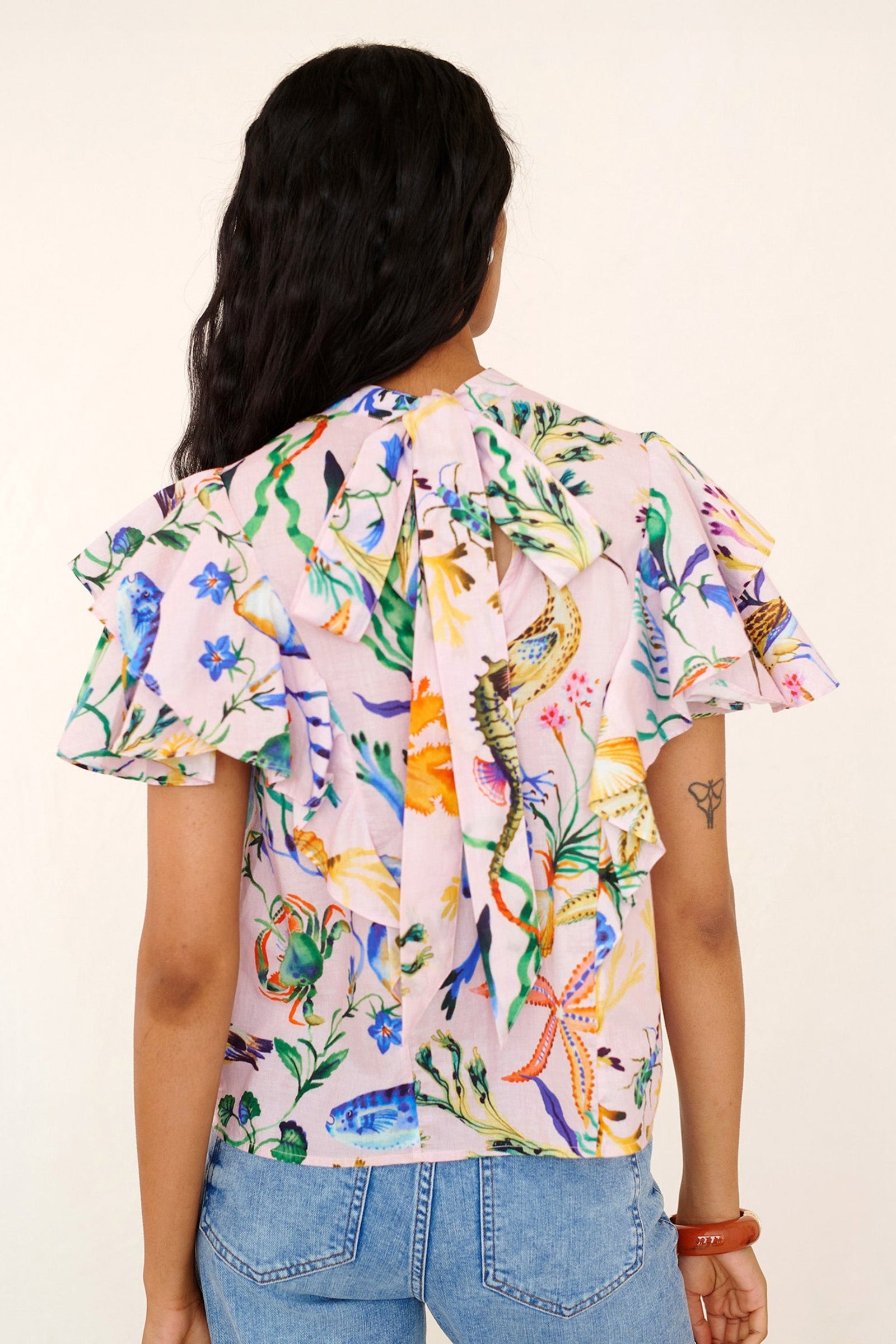 Back view of a woman standing. She is wearing the Bebe top in Tidepool print. It features a soft drape over the shoulder, tie at the neck, and full layered sleeves.