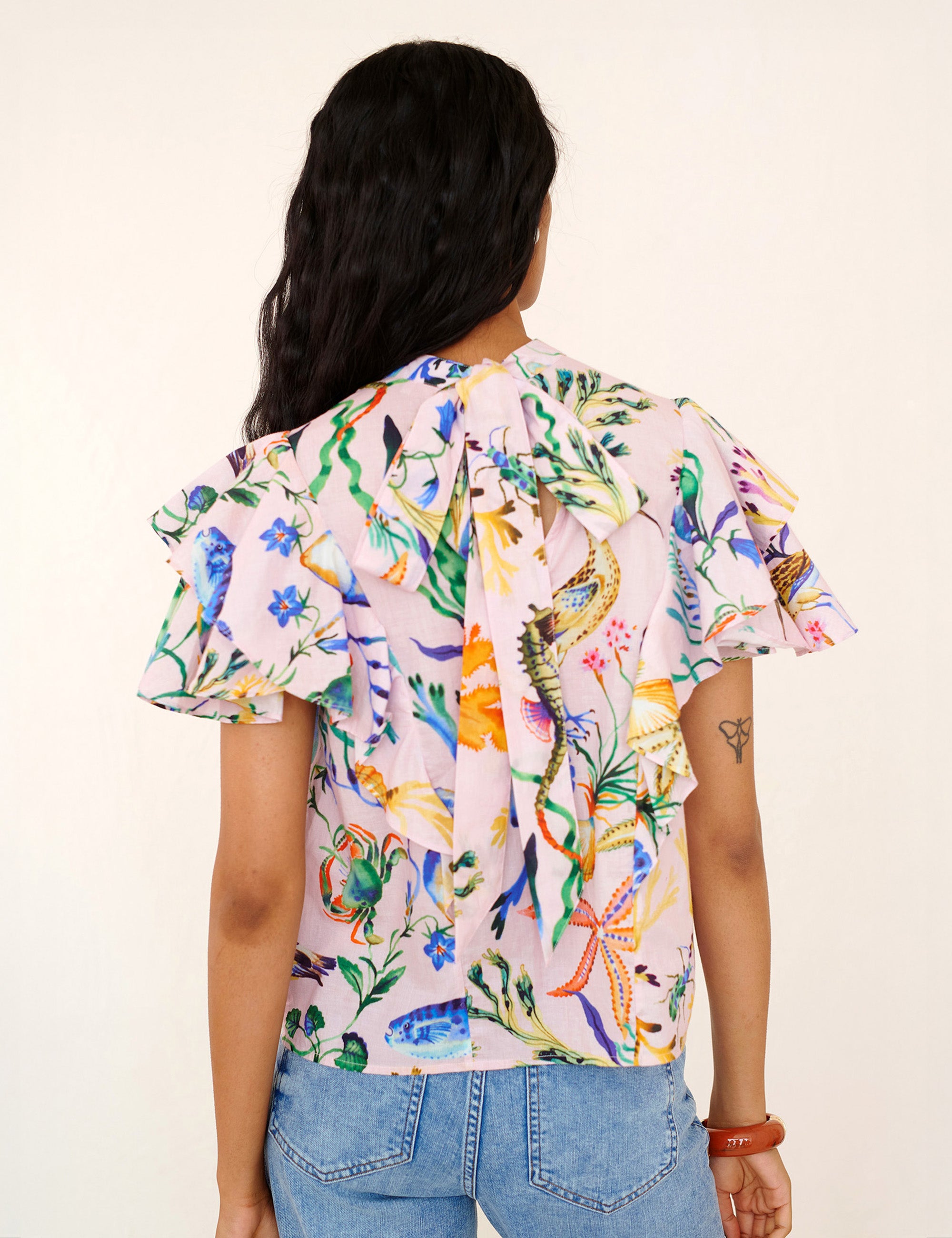 Back view of a woman standing. She is wearing the Bebe top in Tidepool print. It features a soft drape over the shoulder, tie at the neck, and full layered sleeves.