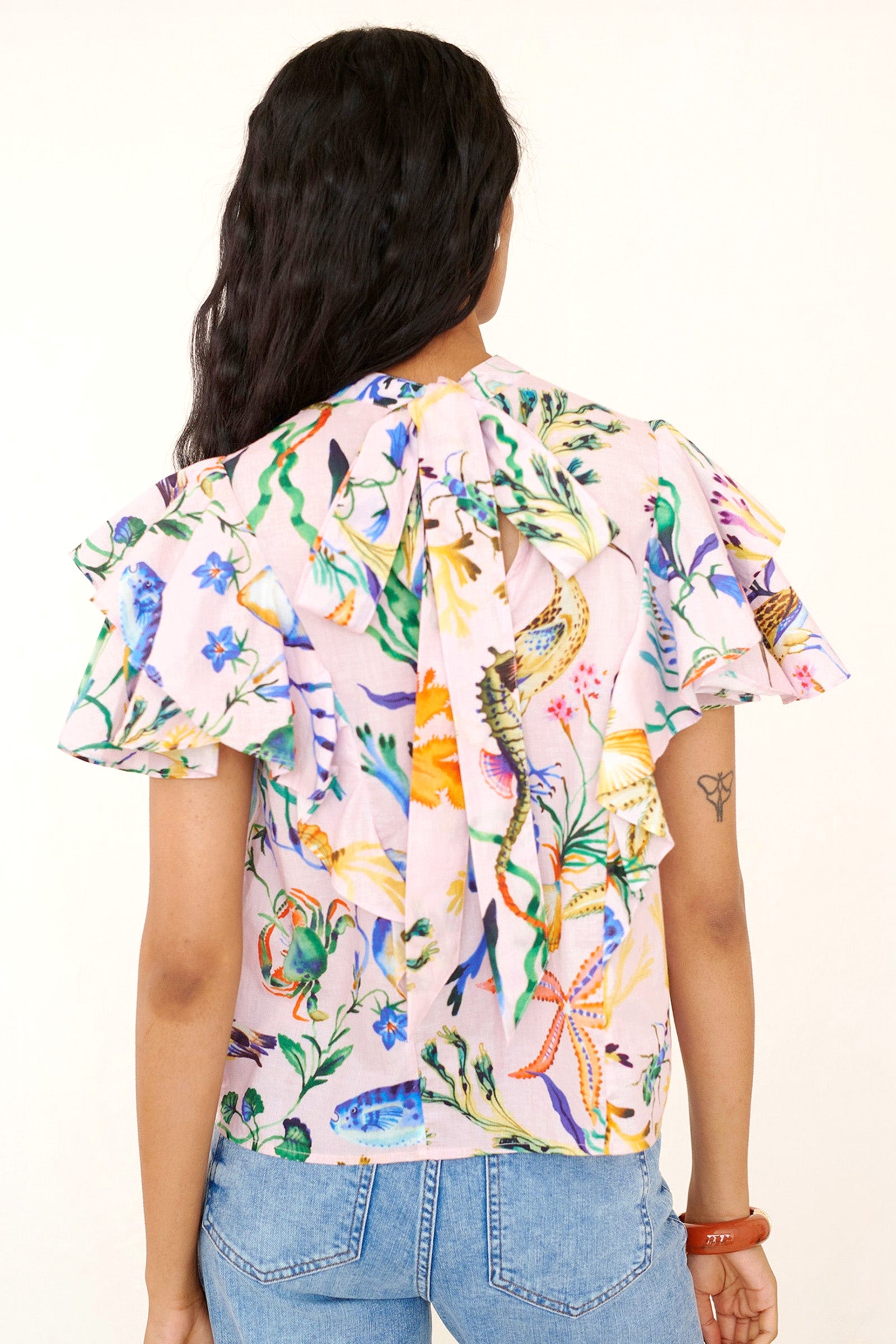 Back view of a woman standing wearing the Bebe top in Tidepool print, featuring a soft shoulder drape, tie neckline, and full layered sleeves. Ocean-inspired print, styled with denim.