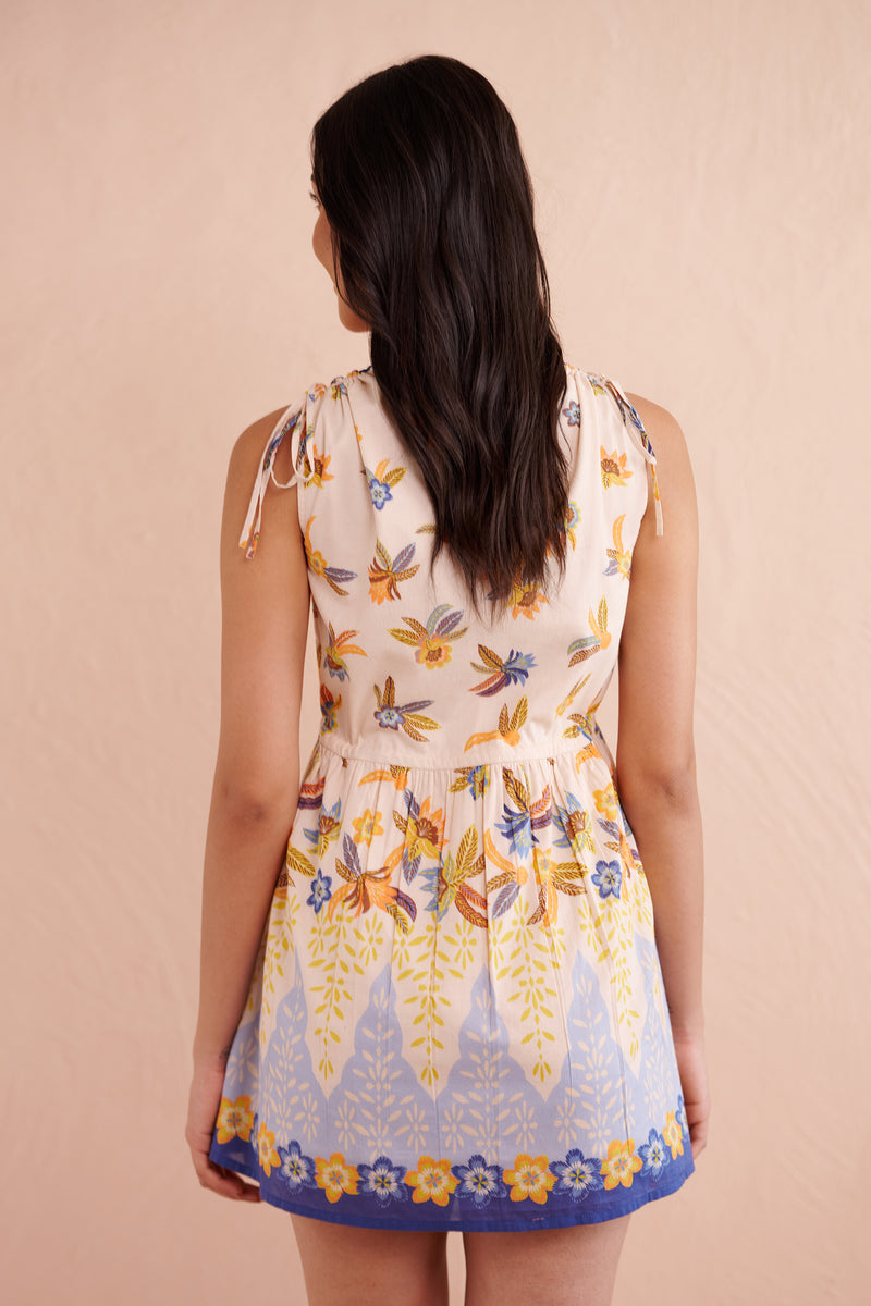 Back view close up of a woman wearing a sleeveless mini dress with a floral and leaf print, featuring a V-neckline with blue floral trim and a gathered drawstring waist.