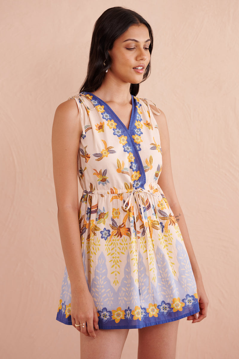 Close up of front view of a woman wearing a sleeveless mini dress with a floral and leaf print, featuring a V-neckline with blue floral trim and a gathered drawstring waist.