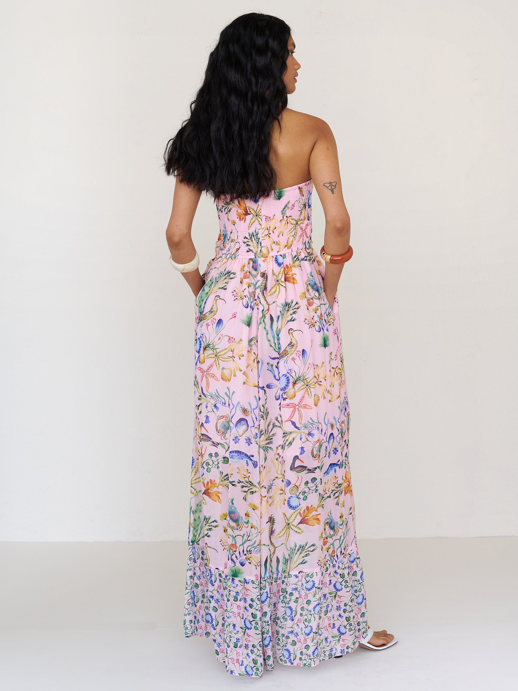 Back view of a woman standing with hands in pocket. She is wearing the Bella full-length dress in Tidepool print. It features a cascading overlapping skirt with frill detail in contrast conch print, ruched bodice, smocked back, and side-seam pockets.