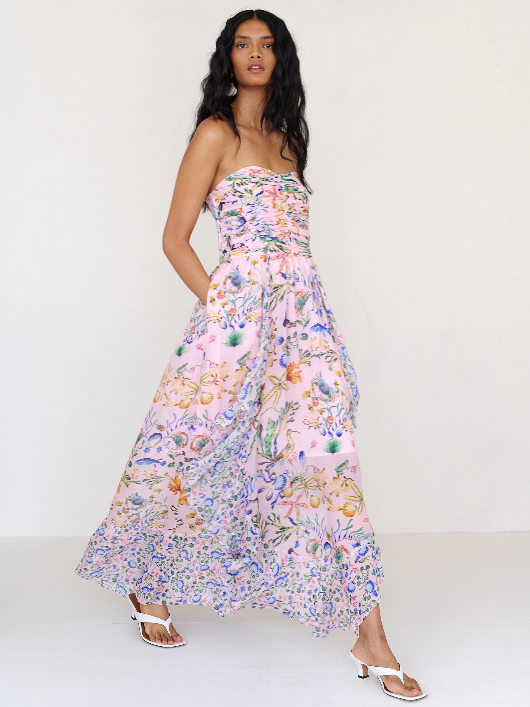 Side view of a woman walking with hands in pocket. She is wearing the Bella full-length dress in Tidepool print. It features a cascading overlapping skirt with frill detail in contrast conch print, ruched bodice, smocked back, and side-seam pockets.