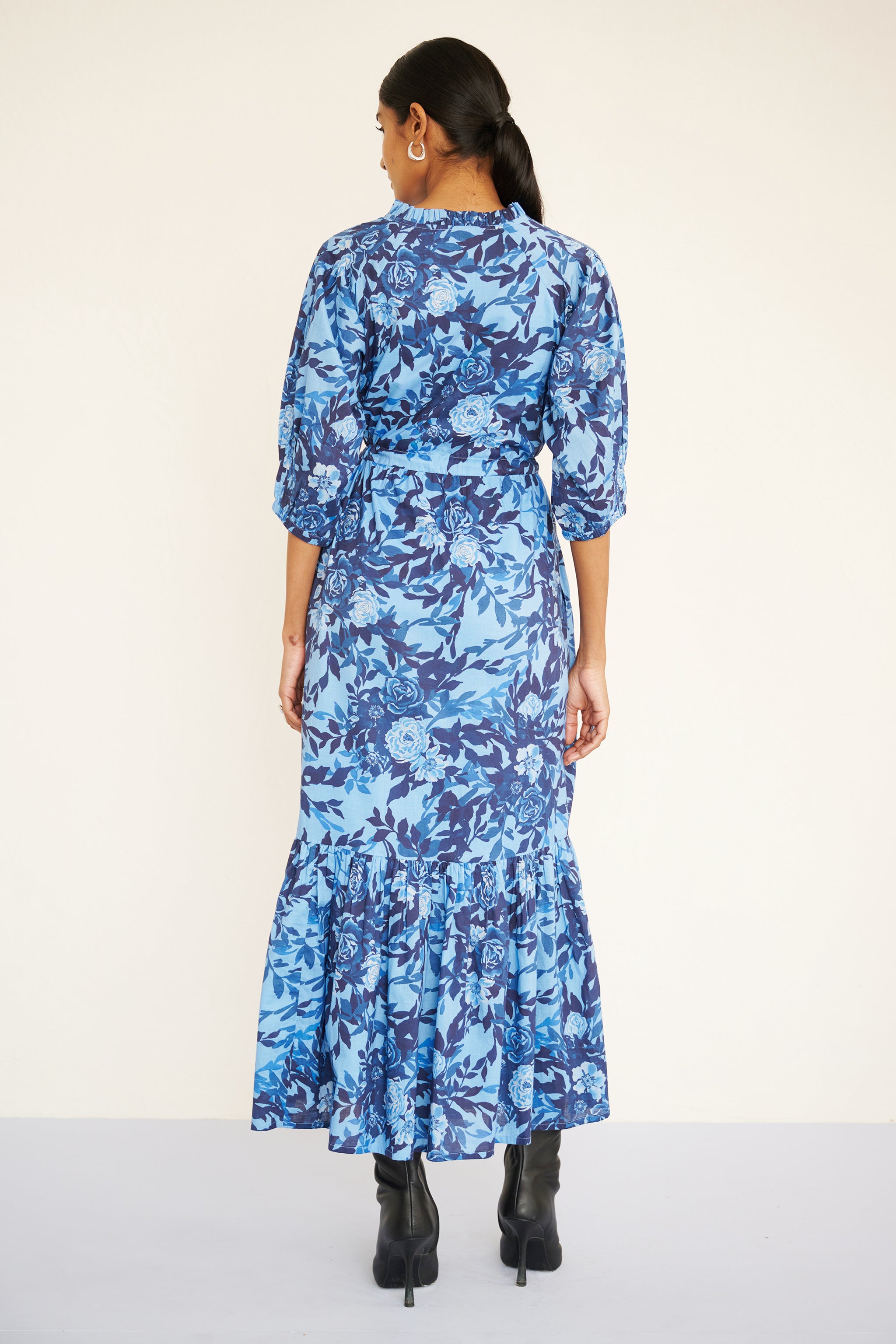 Back view of woman wearing a floral midi dress with raglan sleeves, gathered skirt, tie belt at the waist, and a soft watercolor rose print in tones of blue.