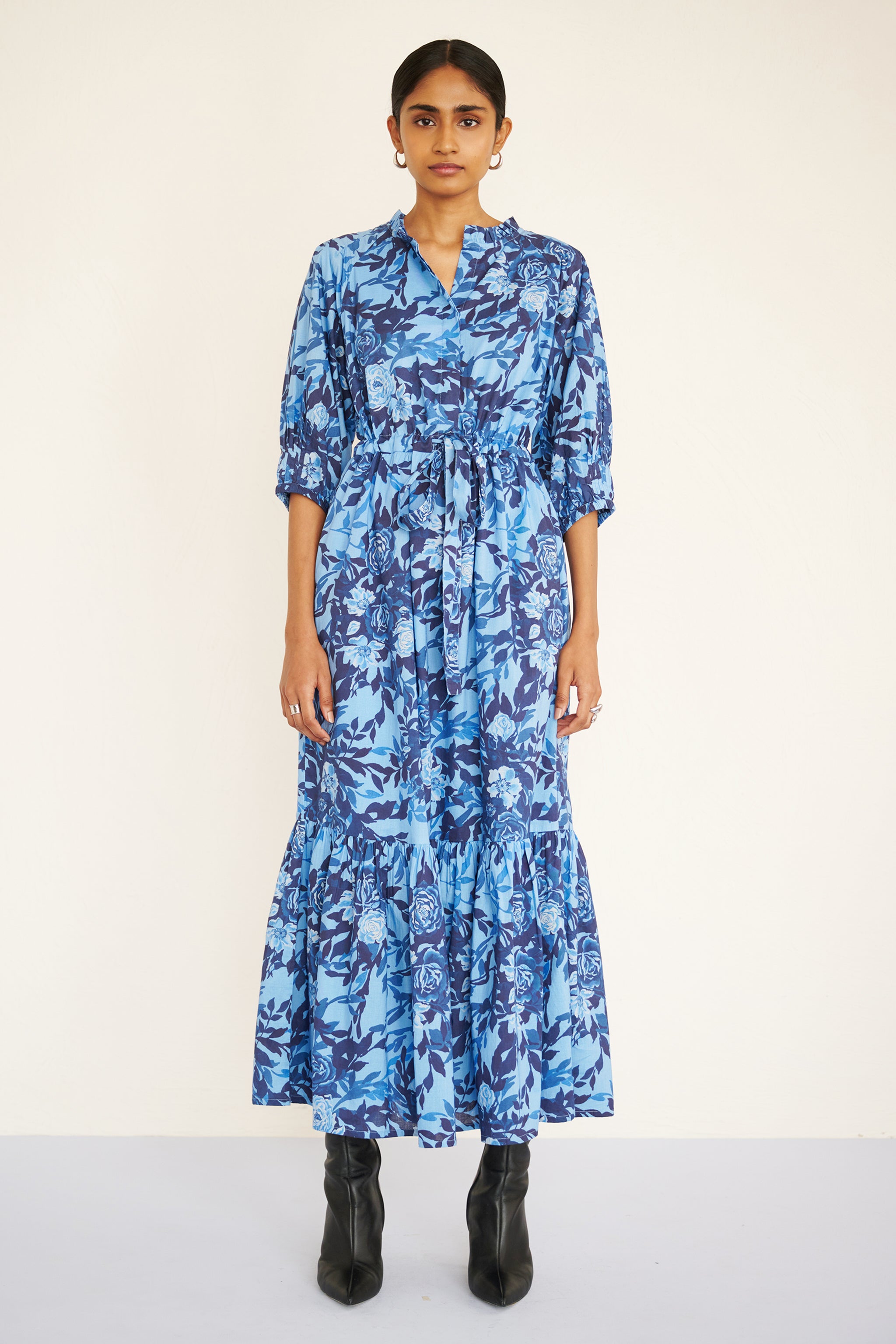 Front view of woman wearing a floral midi dress with raglan sleeves, gathered skirt, tie belt at the waist, and a soft watercolor rose print in tones of blue.