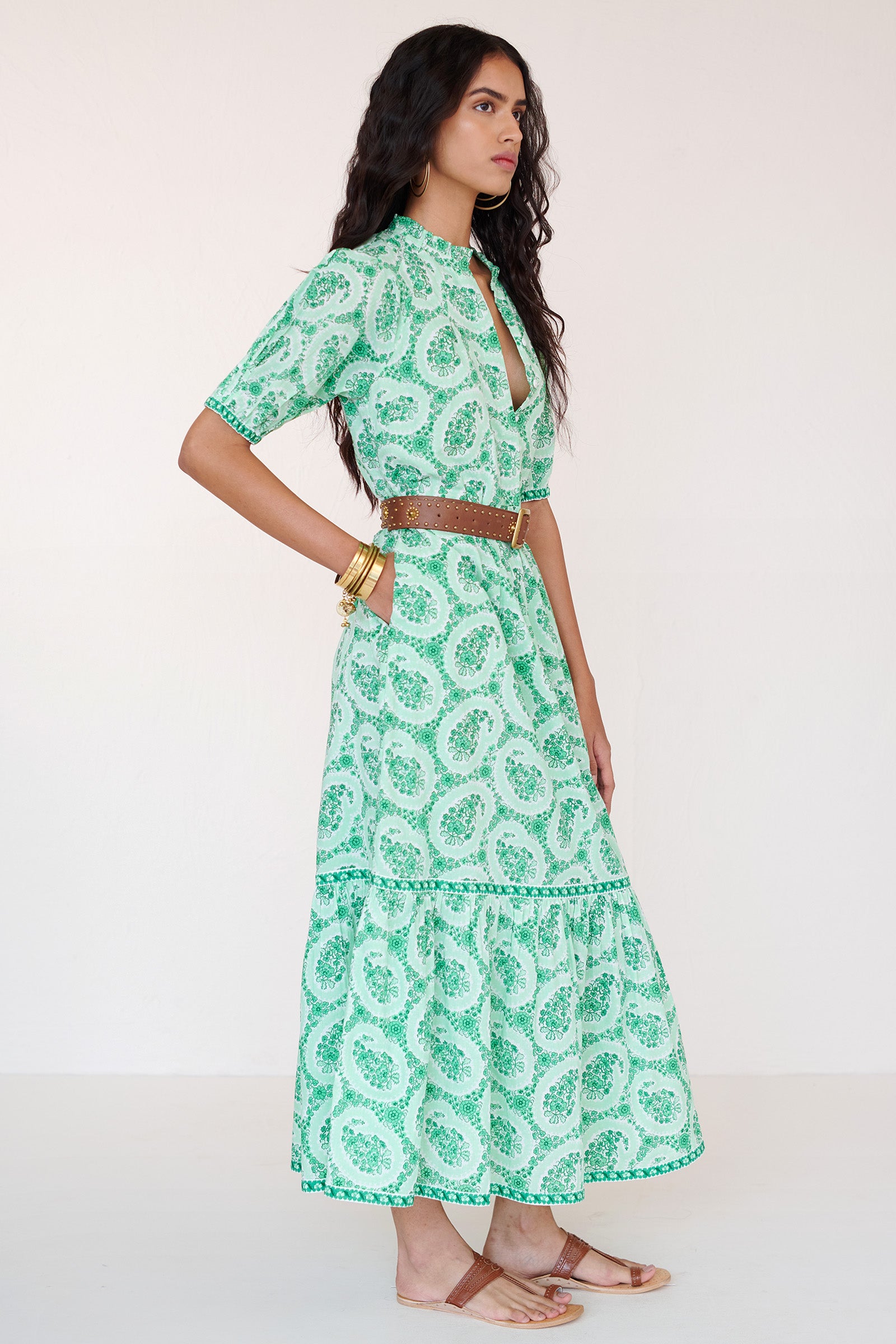 Side view of a woman standing wearing the Betty dress in Paisley Copse print. It features gathered skirt, raglan sleeves, contrast trim and side-seam pockets in breezy cotton voile. Styled with brown belt and gold accessories.