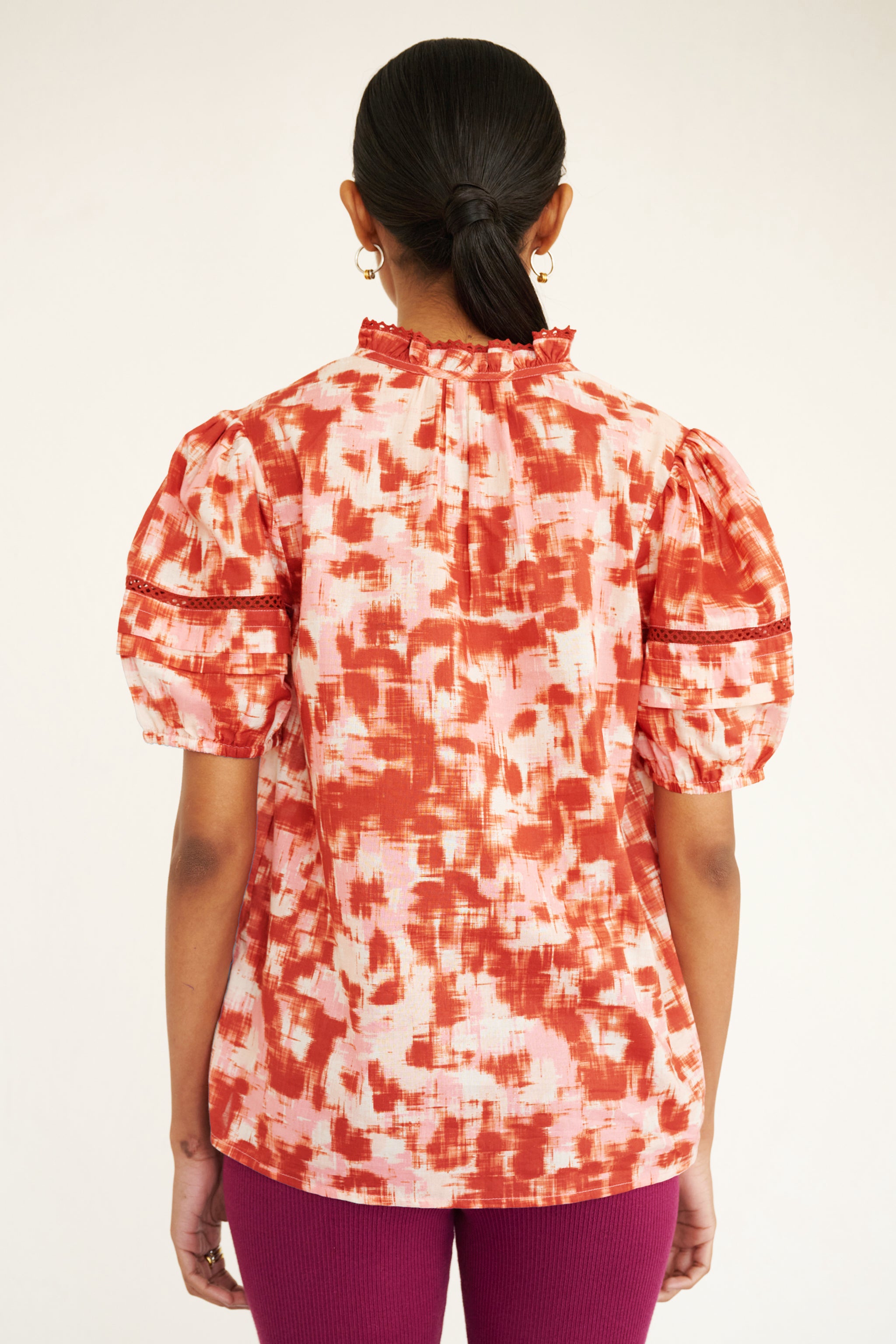 Back view of woman wearing a cotton top with an ikat-inspired print, lace sleeve insets, a split neckline, and pleated sleeves.
