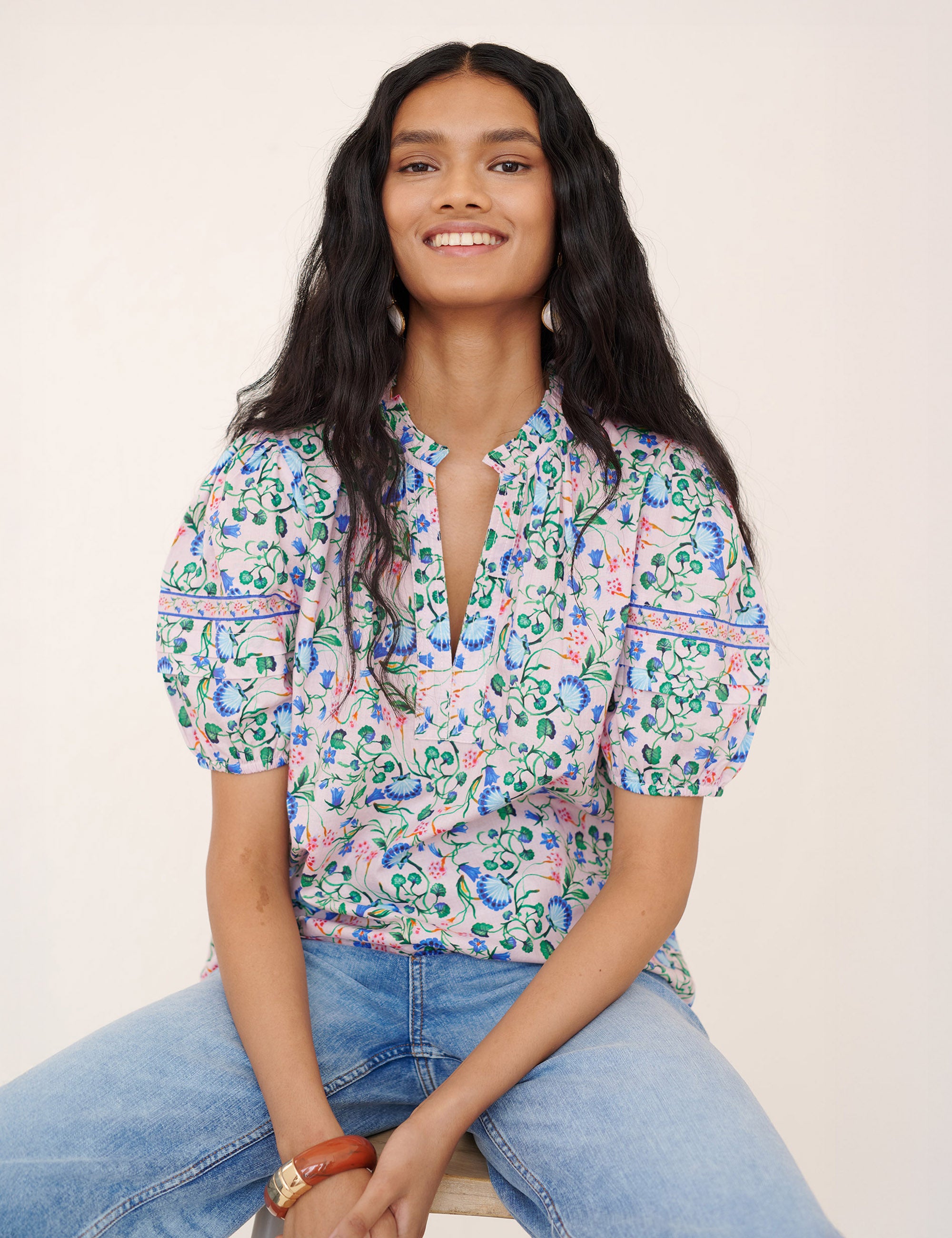Front view of a woman sitting and smiling. She is wearing the Bronte pullover top in Clam Garland print paired with jeans. It features a front opening, contrast trim at the neck, and pleated sleeves.
