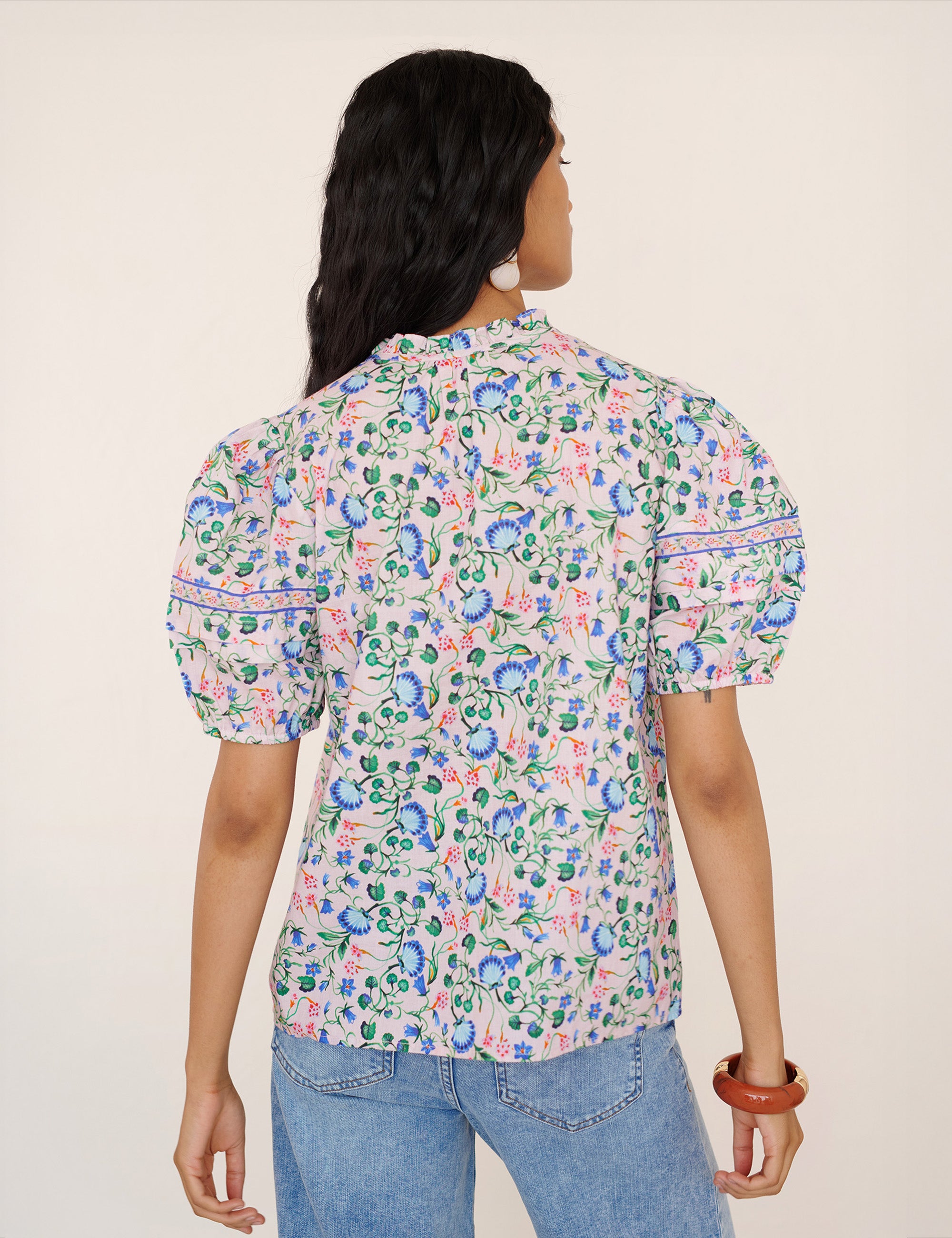 Back view of a woman standing. She is wearing the Bronte pullover top in Clam Garland print paired with jeans. It features a front opening, contrast trim at the neck, and pleated sleeves.