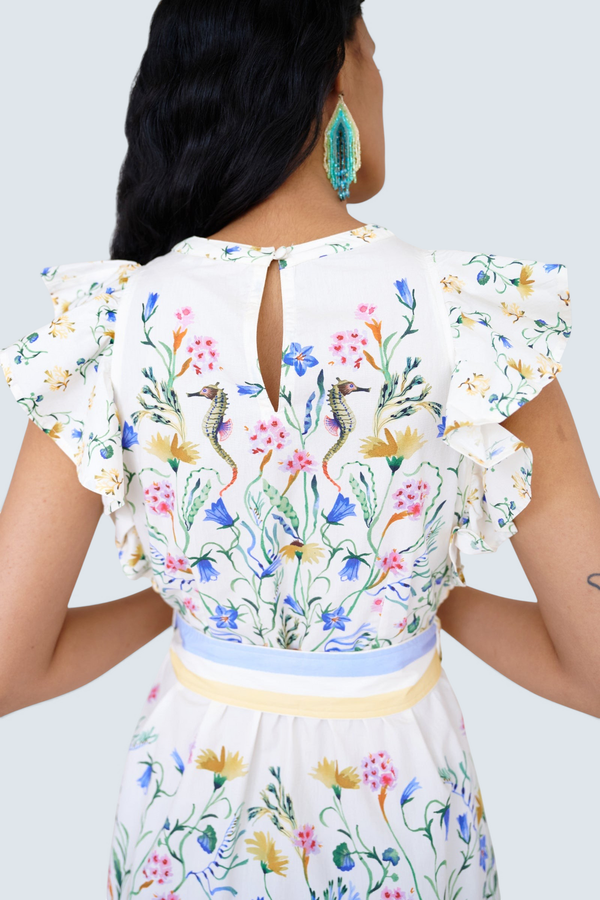Back view close up of a woman standing. She is wearing the Bulbul dress in Tidepool print. It features ruffled shoulders, removable belt, keyhole back, side-seam pockets.