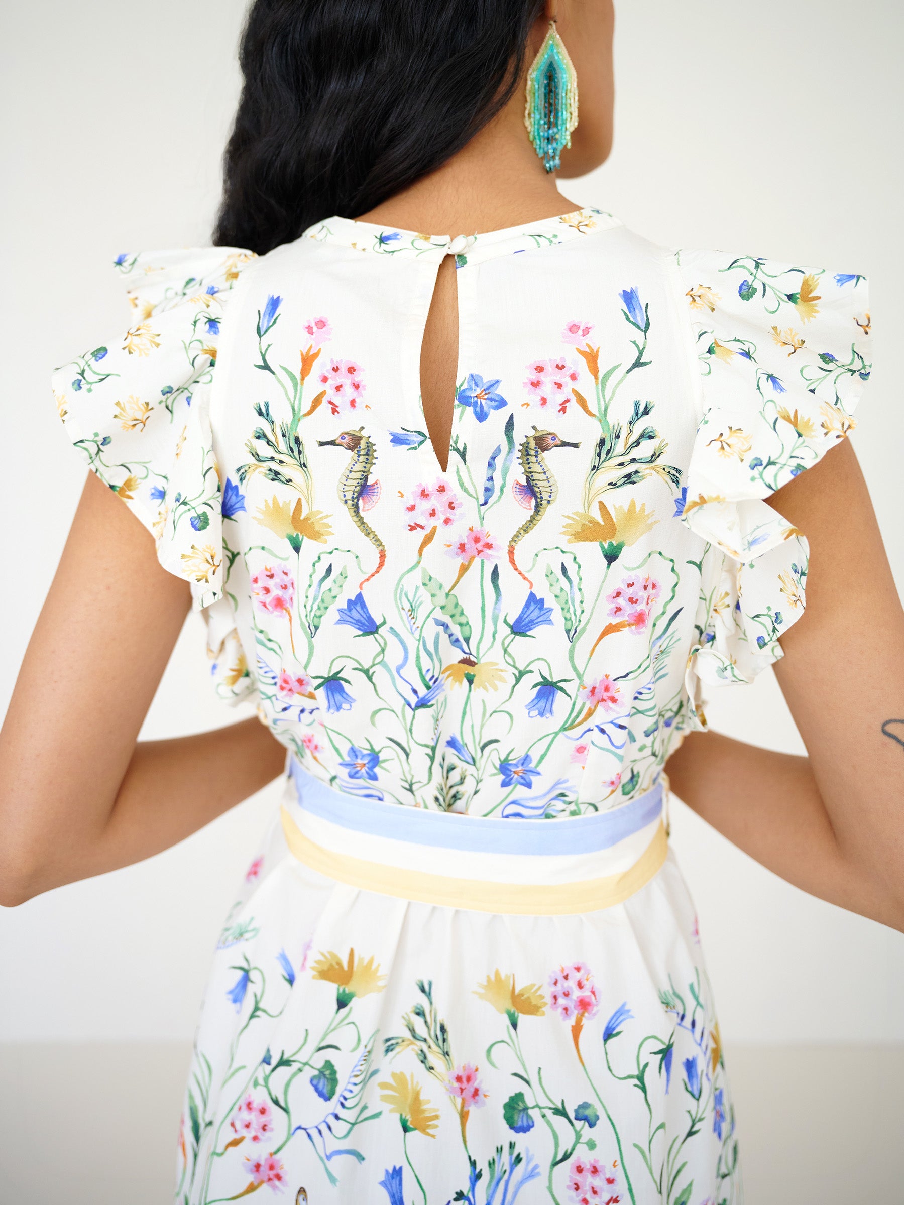 Back view close-up of a woman standing, wearing the Bulbul dress in Tidepool print, showing ruffled shoulders and motifs of seaside wildflowers, seahorses, and oyster catchers.