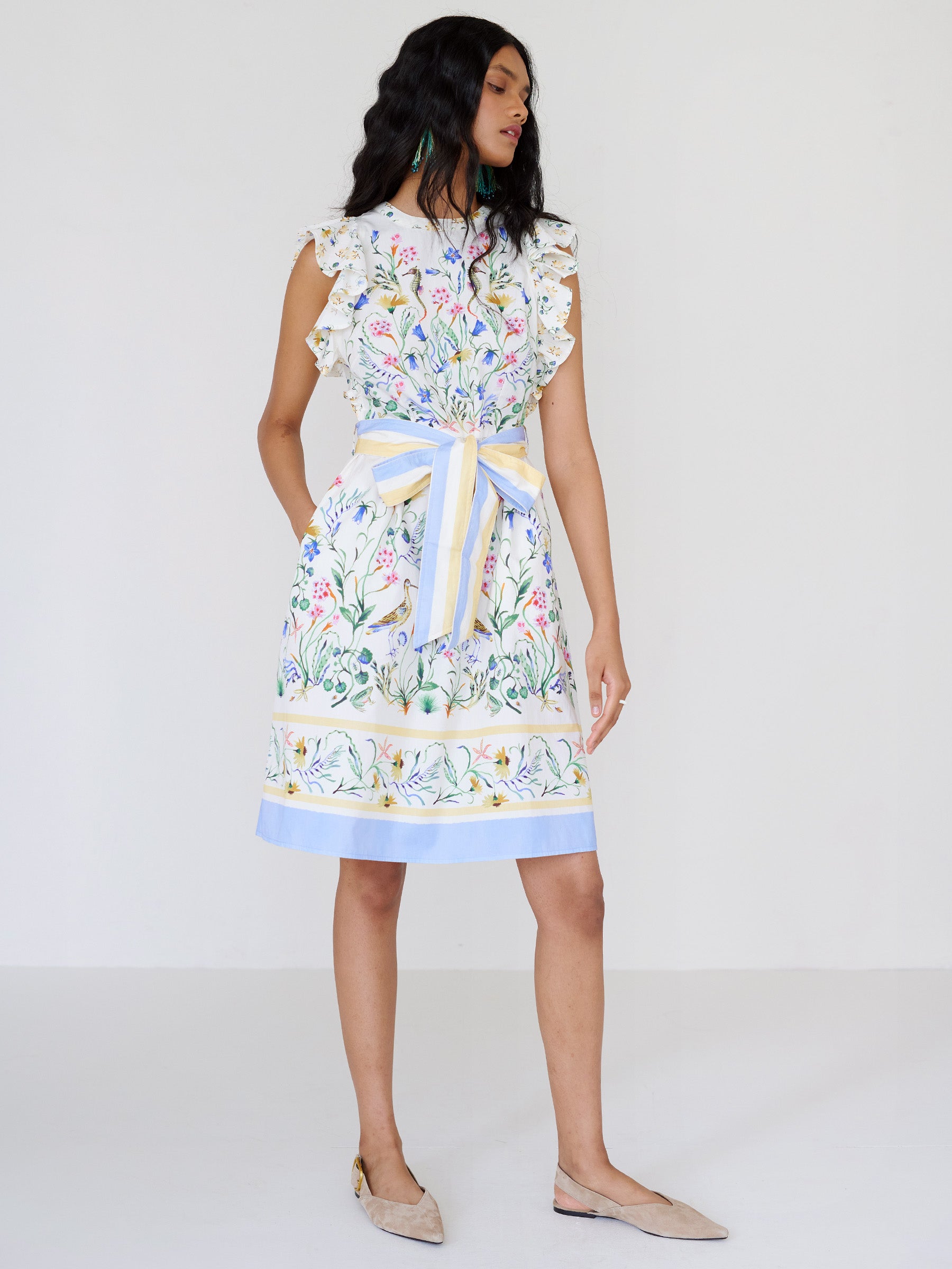 Front view of a woman standing looking sideways. She is wearing the Bulbul dress in Tidepool print. It features ruffled shoulders, a soft hourglass silhouette, a removable self-belt with blue and yellow stripe, and side-seam pockets.