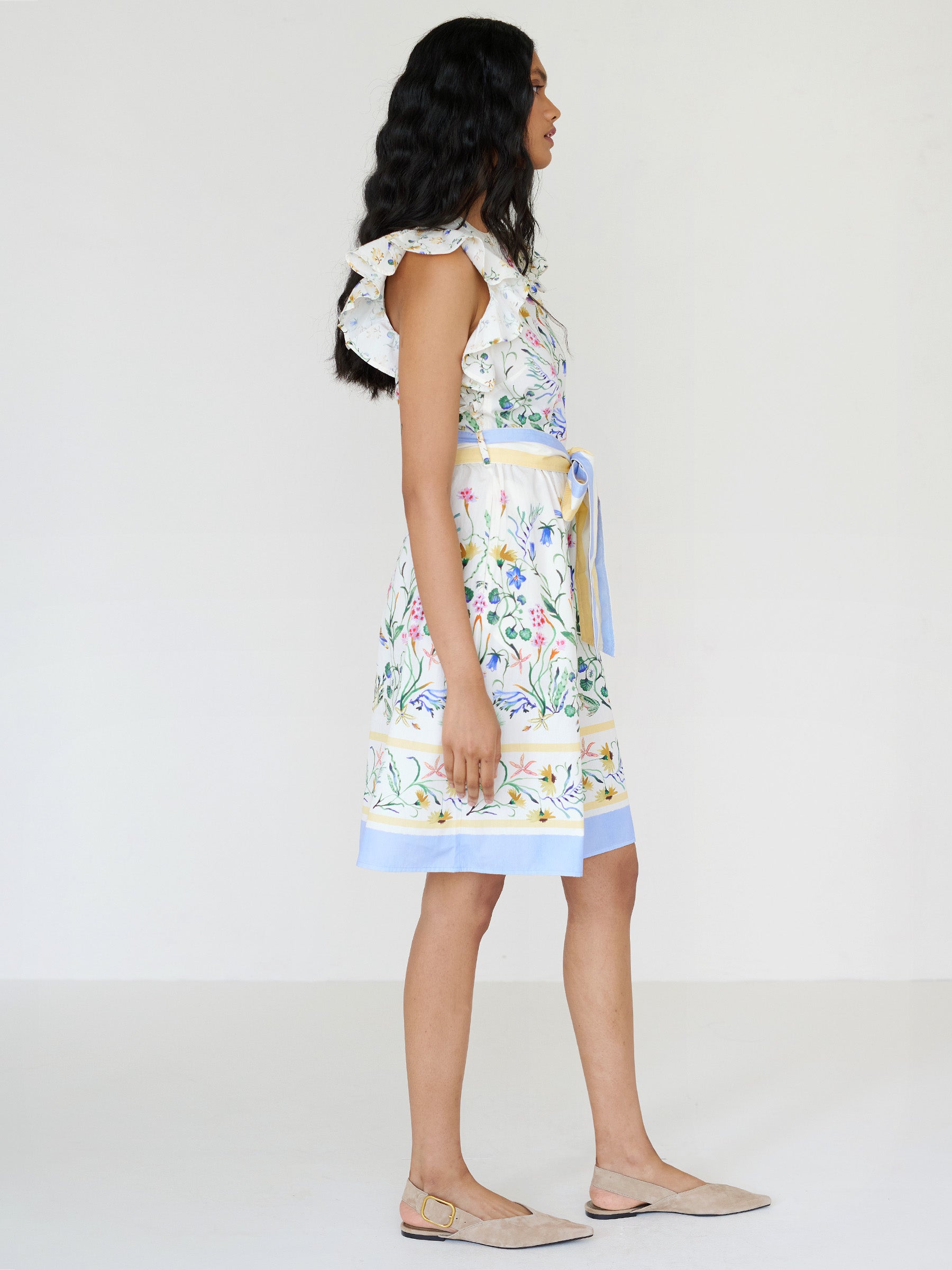 Side view of a woman standing. She is wearing the Bulbul dress in Tidepool print. It features ruffled shoulders, a soft hourglass silhouette, a removable self-belt with blue and yellow stripe, and side-seam pockets.