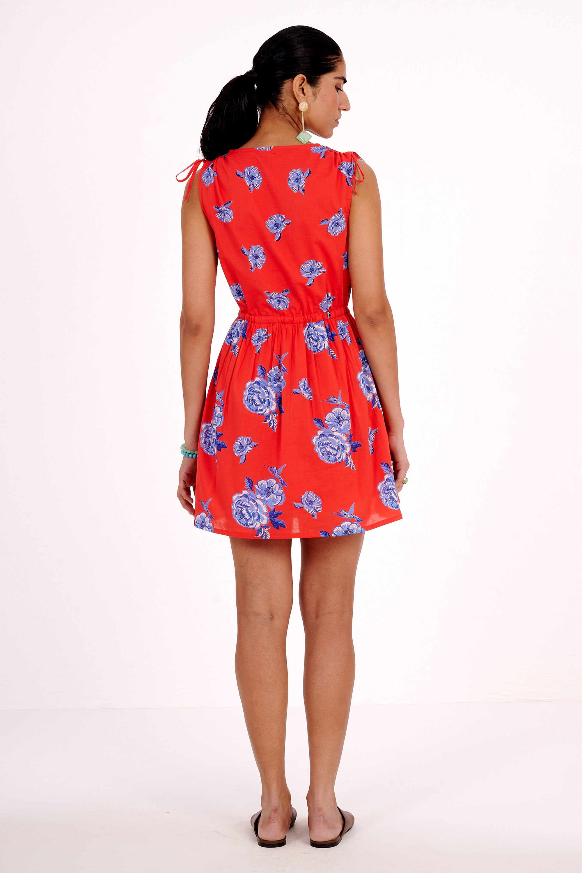 Back view of woman standing. She is  wearing the Becca dress in the Eliza Rose print, featuring a mini-dress silhouette, adjustable waist, shoulder ties, and floral motifs with blue roses and hummingbirds.