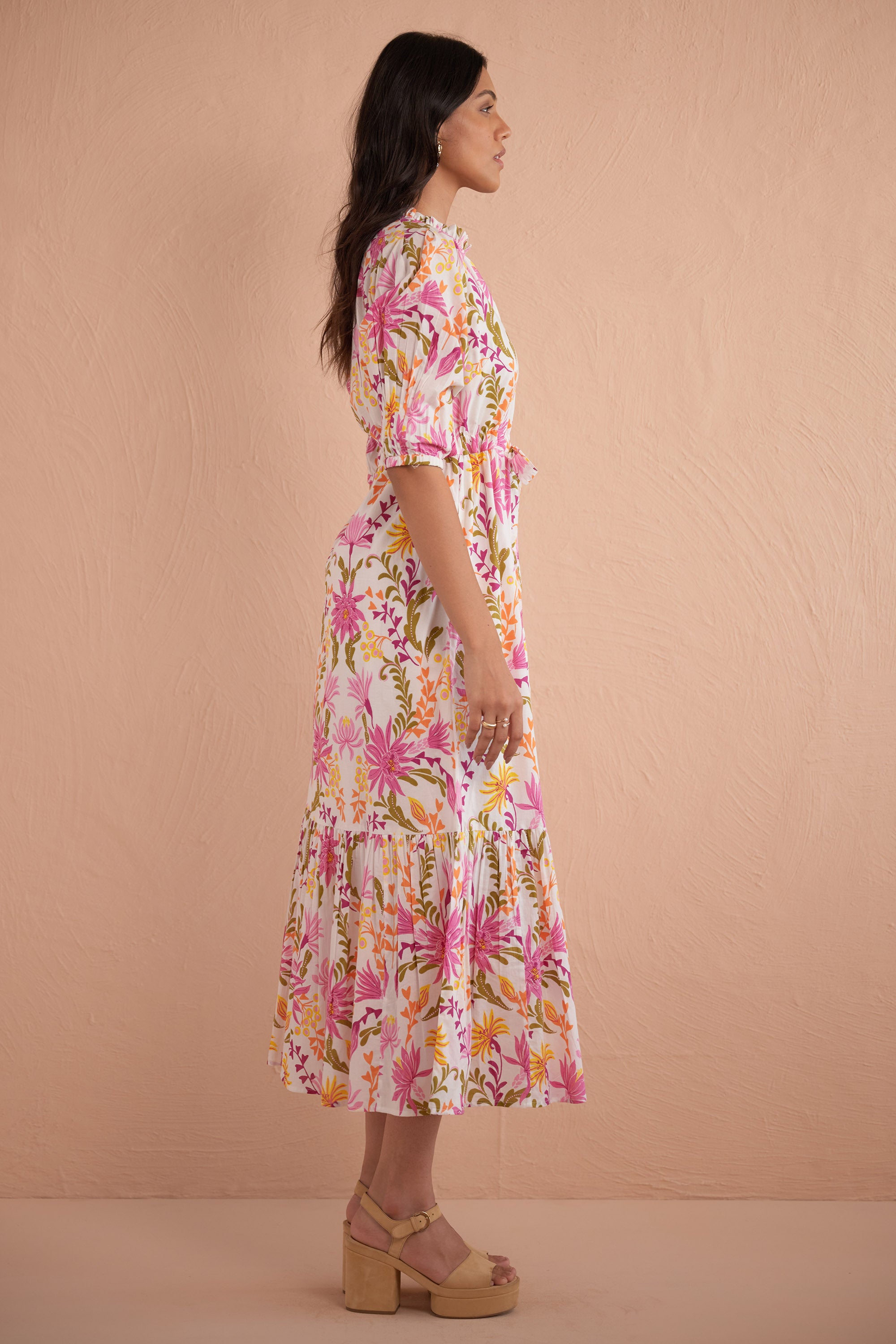 Side view of woman wearing Betty Dress in Hydra Garland Rose print, featuring a cinched waist, full gathered skirt, tucked raglan sleeves, concealed front button placket, drawstring waist, and side-seam pockets in bold sunset floral cotton voile.