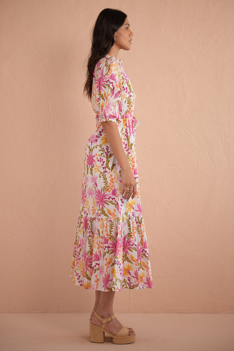 Side view of woman wearing Betty Dress in Hydra Garland Rose print, featuring a cinched waist, full gathered skirt, tucked raglan sleeves, concealed front button placket, drawstring waist, and side-seam pockets in bold sunset floral cotton voile.