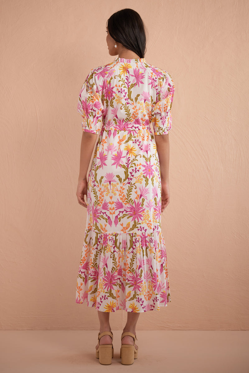 Back view of woman wearing Betty Dress in Hydra Garland Rose print, featuring a cinched waist, full gathered skirt, tucked raglan sleeves, concealed front button placket, drawstring waist, and side-seam pockets in bold sunset floral cotton voile.