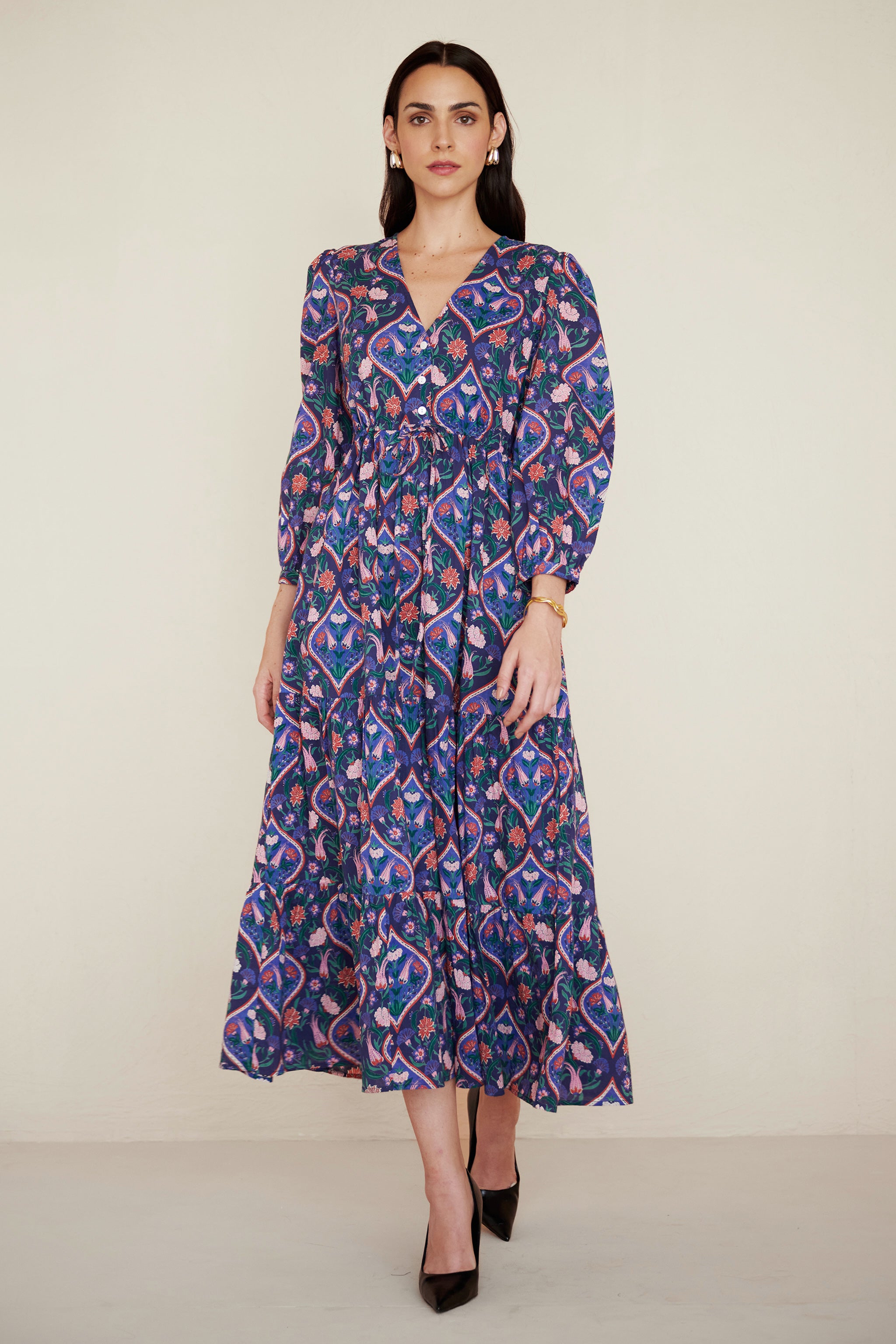Front view of woman walking. She is wearing a Castor dress in Persian Tile print in Lapis color. It is a cotton midi dress with puff sleeves and a gathered drawstring skirt, featuring a floral tile print and side pockets.