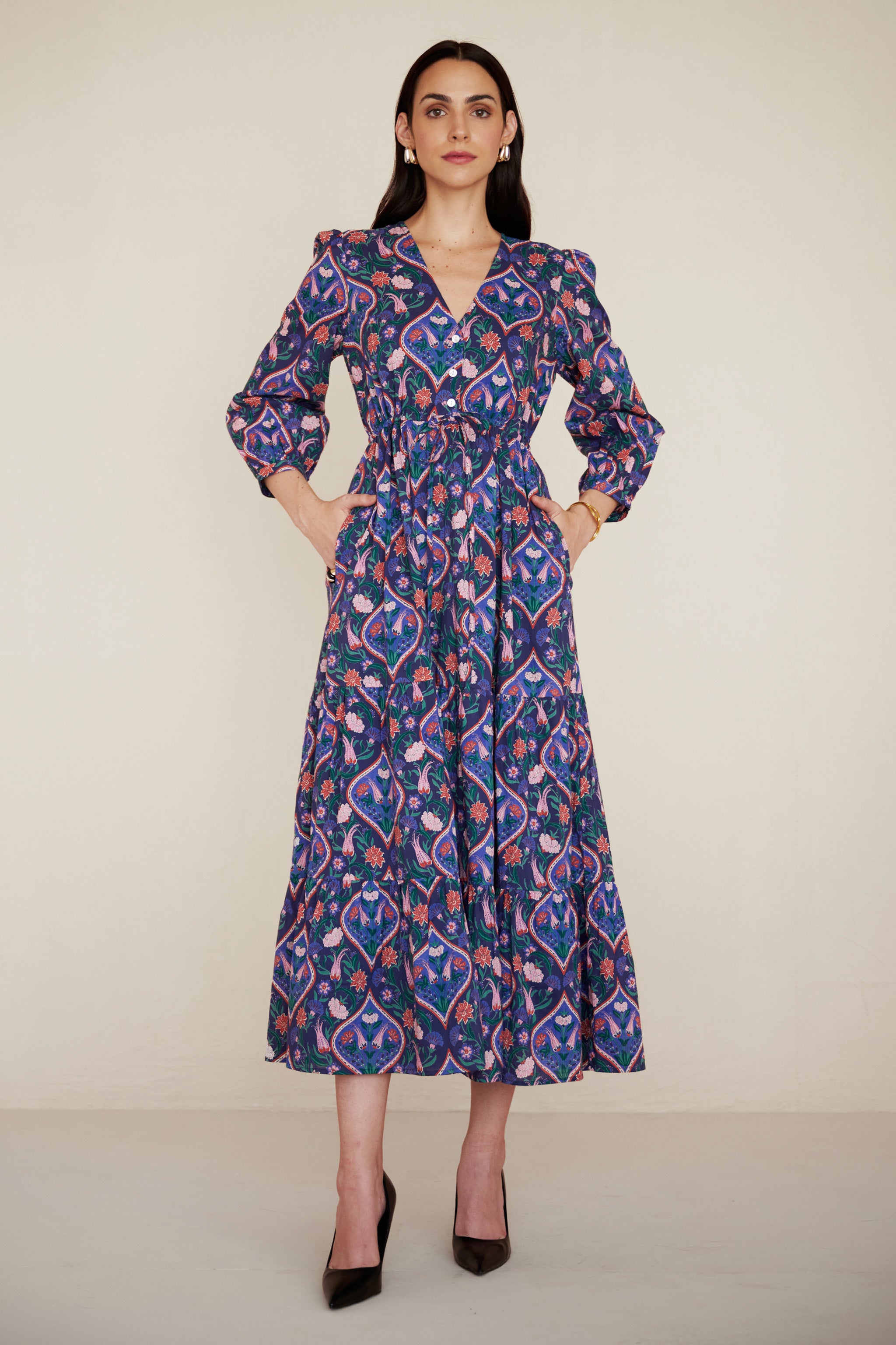 Front view of woman wearing a Castor dress in Persian Tile print in Lapis color with her both hands in pocket. It is a cotton midi dress with puff sleeves and a gathered drawstring skirt, featuring a floral tile print and side pockets.