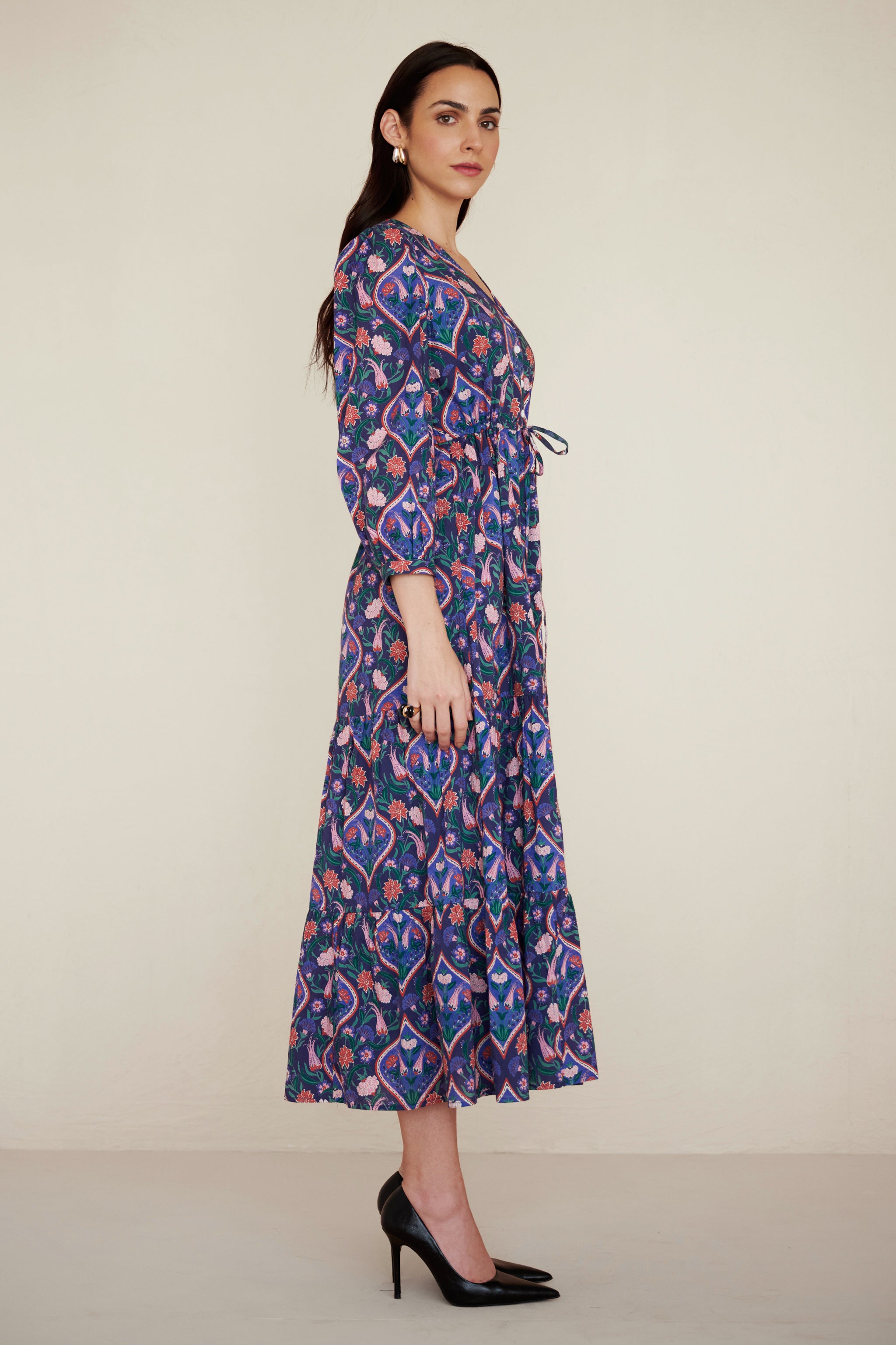 Side view of woman wearing a Castor dress in Persian Tile print in Lapis color. It is a cotton midi dress with puff sleeves and a gathered drawstring skirt, featuring a floral tile print and side pockets.
