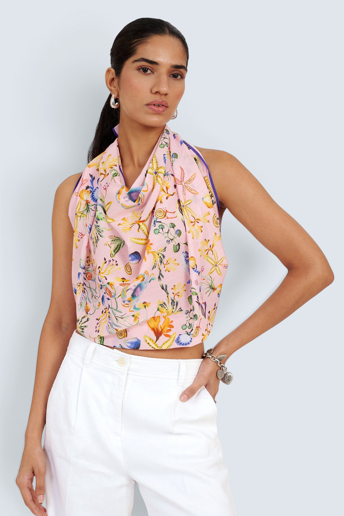 Front view of woman standing. She is wearing the Carolina scarf as a top in the Tidepool print, featuring starfish, seahorses, and shell motifs.Paired with white pants.