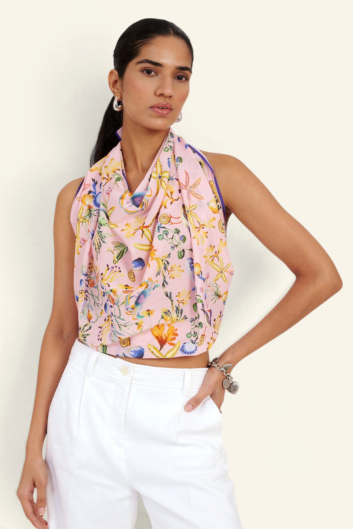 Front view of woman standing. She is wearing the Carolina scarf as a top in the Tidepool print, featuring starfish, seahorses, and shell motifs.Paired with white pants.