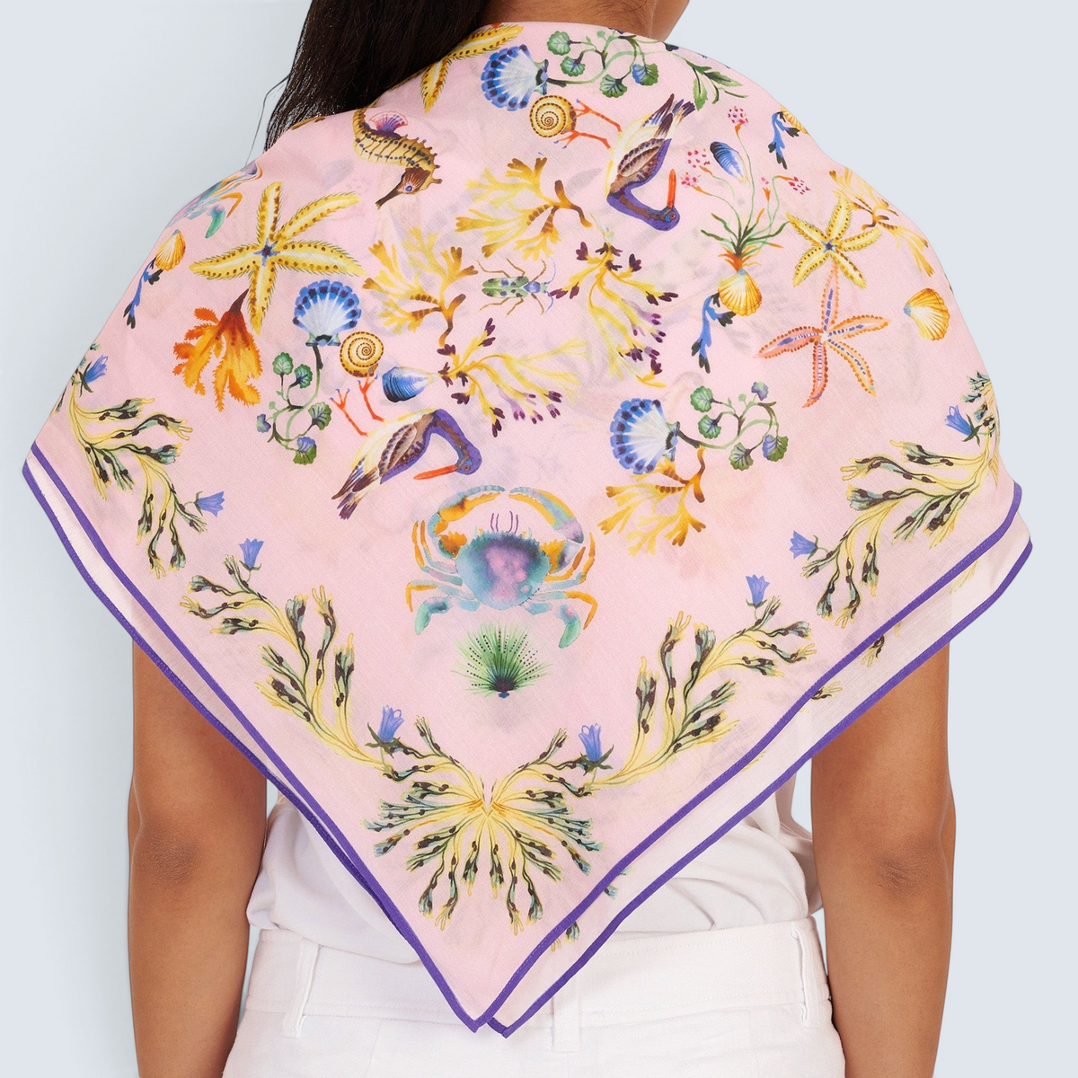 Back view of woman standing, styled with the Carolina scarf draped over her shoulder in the Tidepool print featuring starfish, seahorse, and shell motifs, paired with white pants.