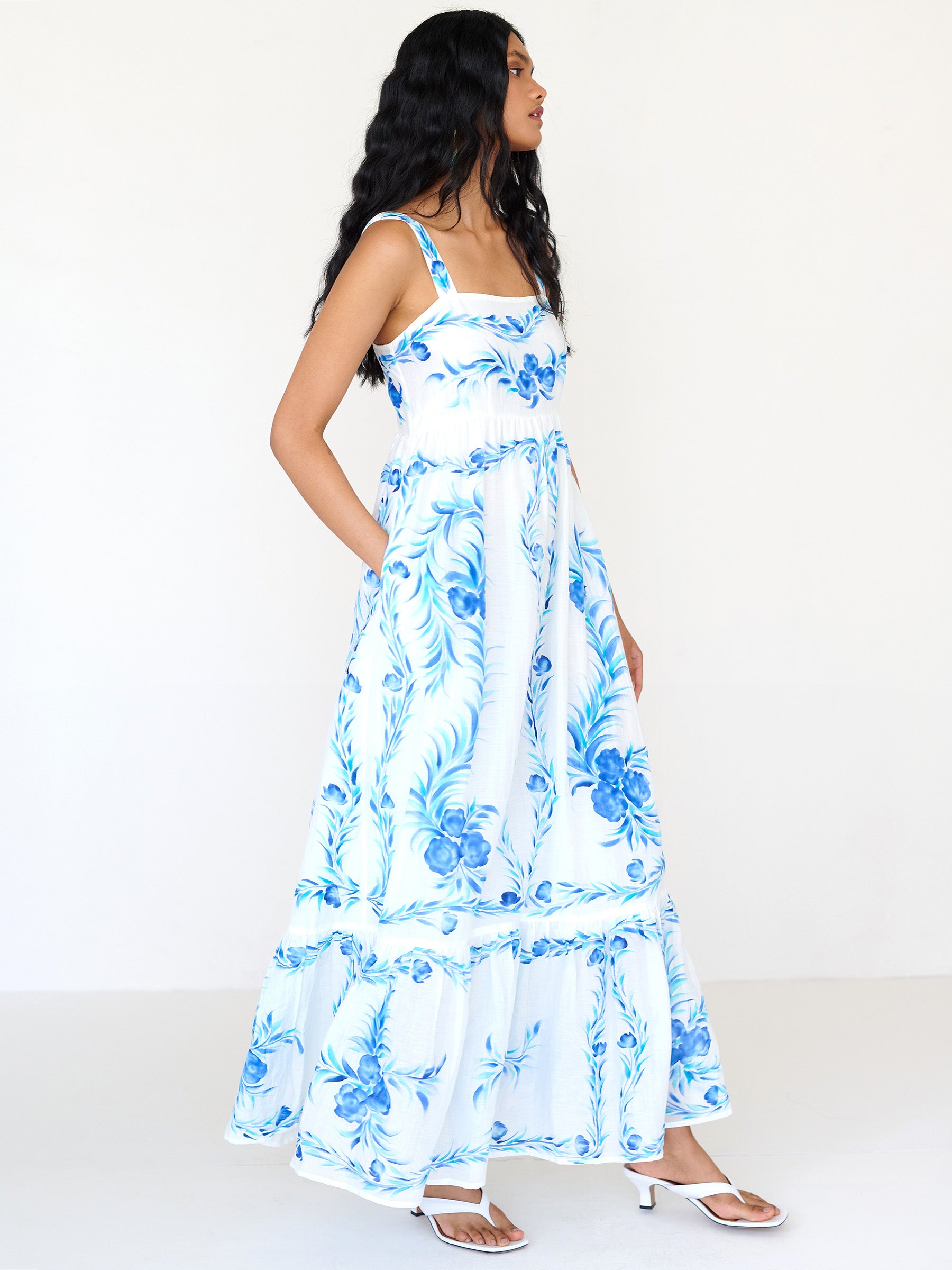 Side view of a woman standing with hand in pocket. She is wearing the Daniella dress with a hand-painted undulating floral print. It features a straight neckline, ankle-grazing hem, and a sweeping silhouette.