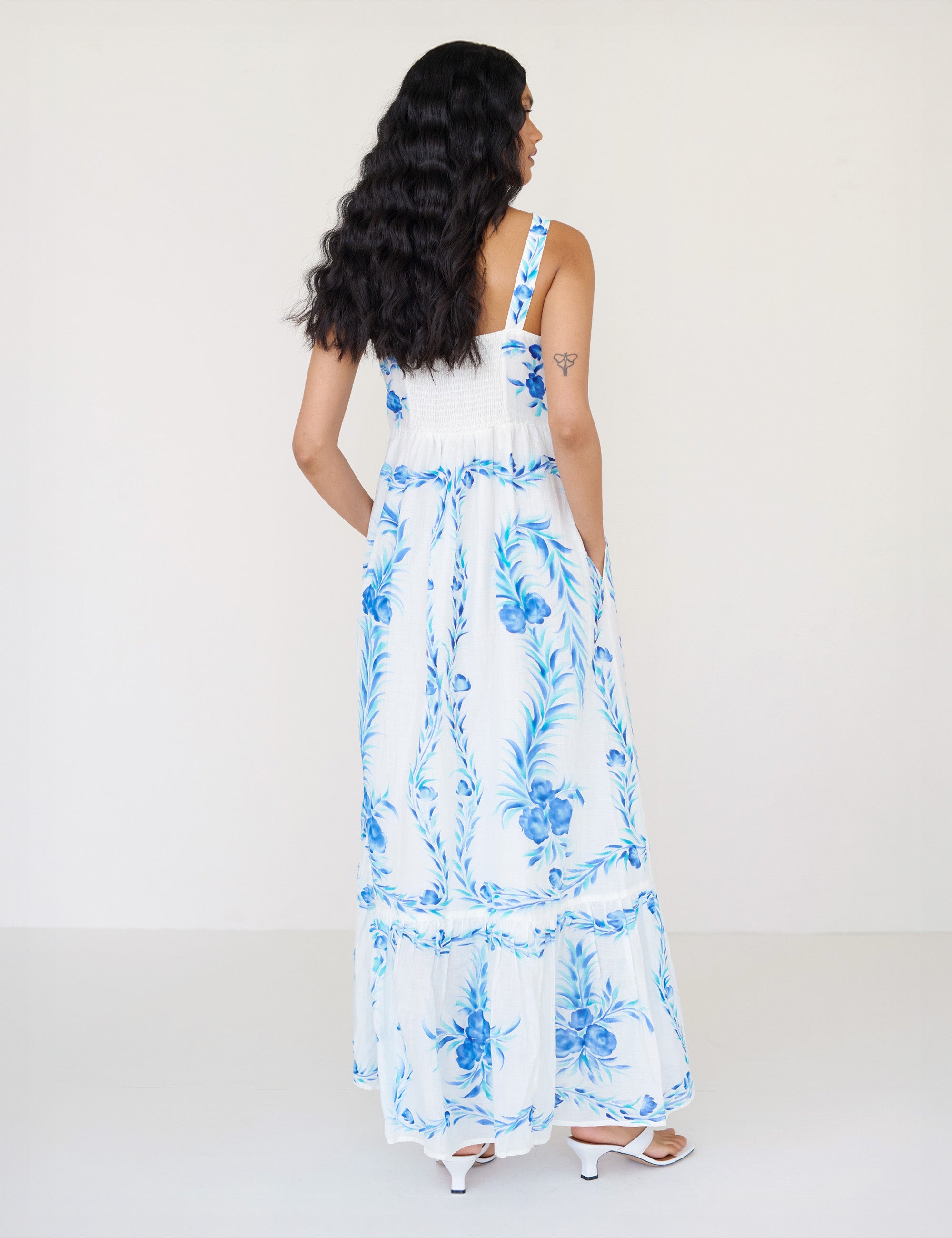 Back view of a woman standing with hands in pocket. She is wearing the Daniella dress with a hand-painted undulating floral print. It features a straight neckline, ankle-grazing hem, and a sweeping silhouette.