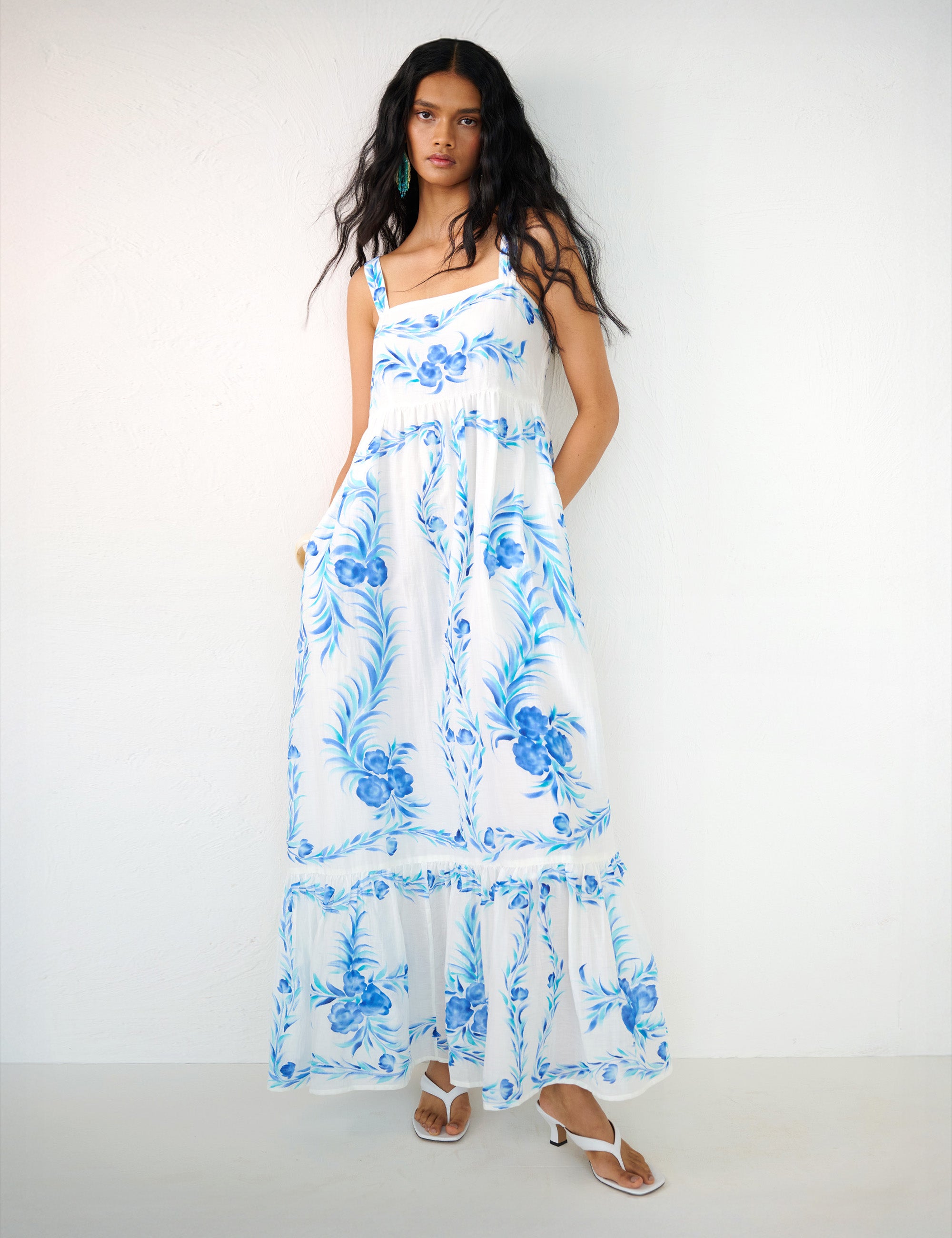 Front view of a woman standing with hands in pocket. She is wearing the Daniella dress with a hand-painted undulating floral print. It features a straight neckline, ankle-grazing hem, and a sweeping silhouette.