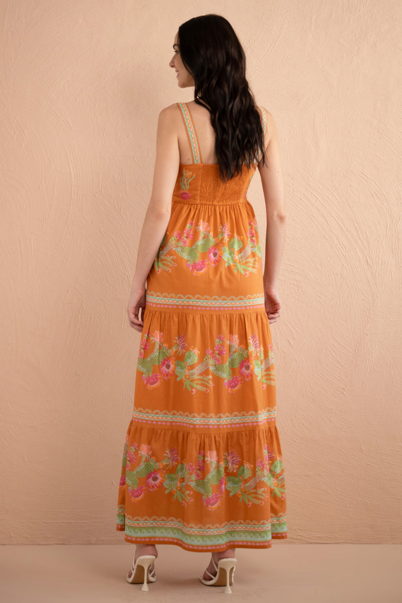 Back view of woman wearing Daniella Dress in Calypso Cacti print, featuring a flowing silhouette, side-zip closure, side-seam pockets, and vibrant cactus blossoms with geometric borders in sun-drenched Aegean colors.