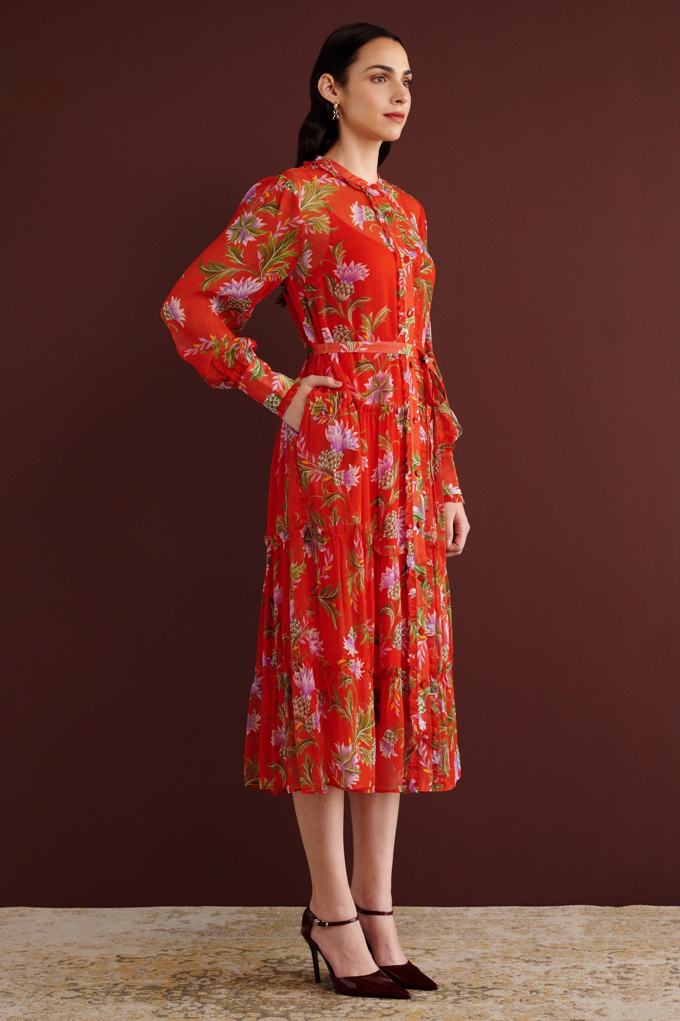 Side view of woman standing with hand in pocket. She is wearing the Emira dress in the Stuart Thistle print, a flowing Flame-colored crinkle chiffon dress with sheer sleeves, delicate ruffles, fabric-covered buttons, and a detachable belt, featuring floral motifs inspired by the Scottish Highlands.