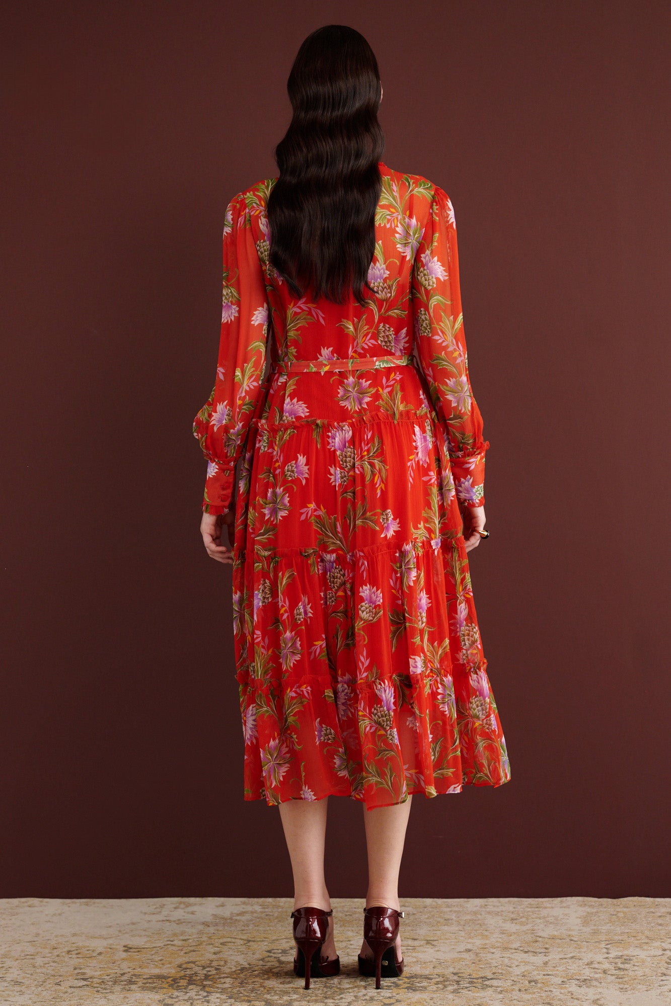 Back view of woman standing. She is wearing the Emira dress in the Stuart Thistle print, a flowing Flame-colored crinkle chiffon dress with sheer sleeves, delicate ruffles, fabric-covered buttons, and a detachable belt, featuring floral motifs inspired by the Scottish Highlands.